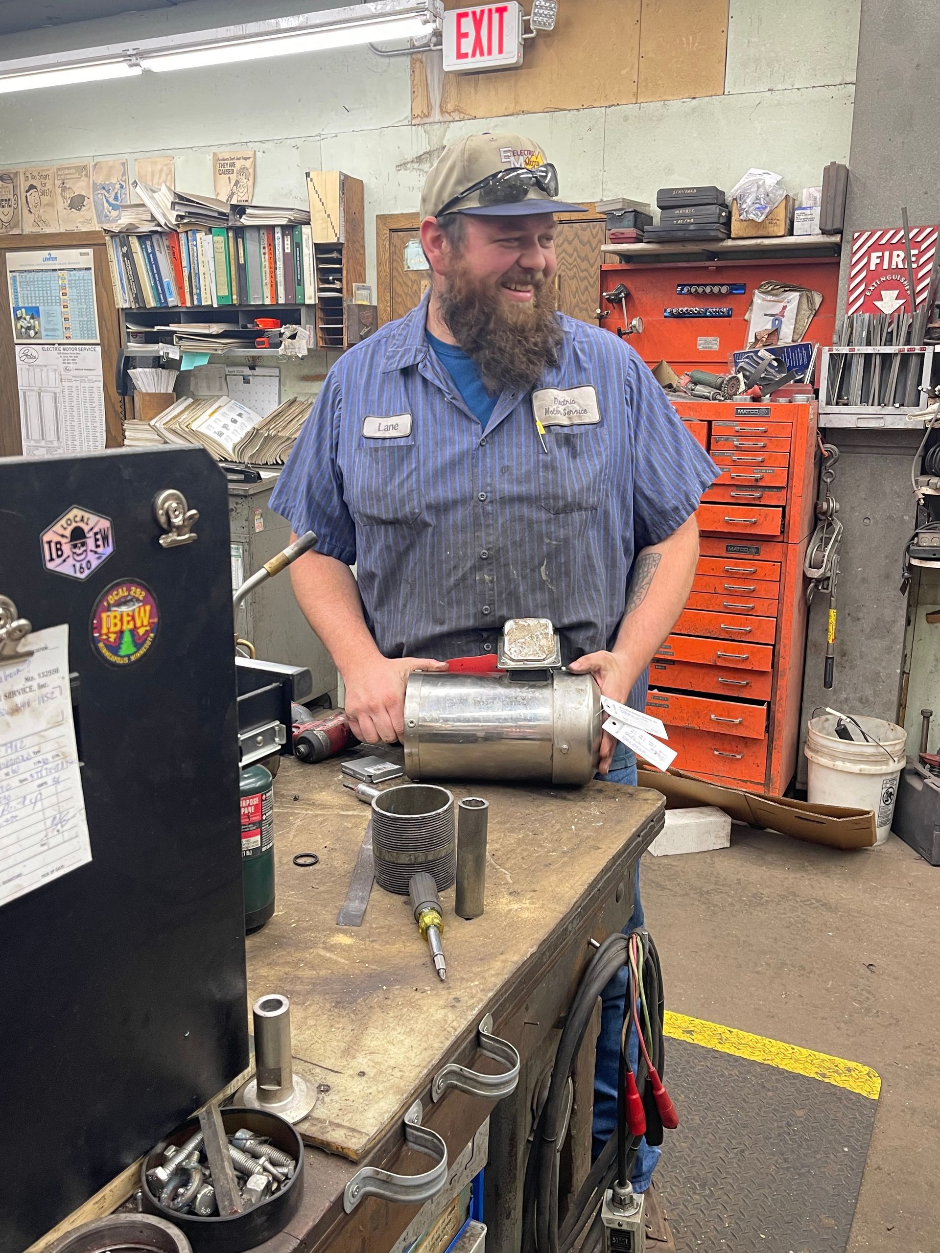 A bearded worker in a blue work shirt smiling while holding a cylindrical motor part at a cluttered workbench.