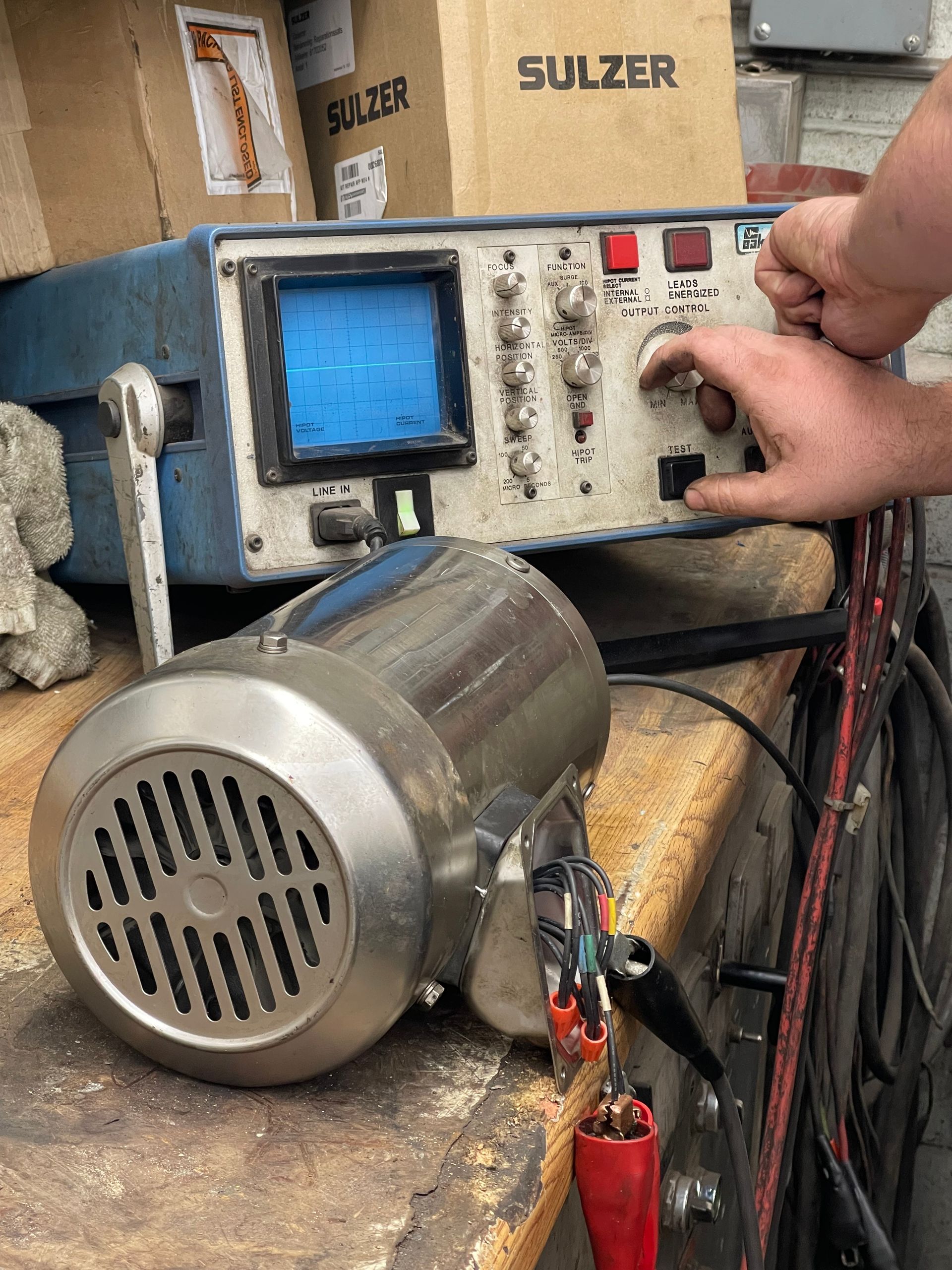 A person's hands adjust a control knob on an oscilloscope connected to a metal electric motor on a workbench.