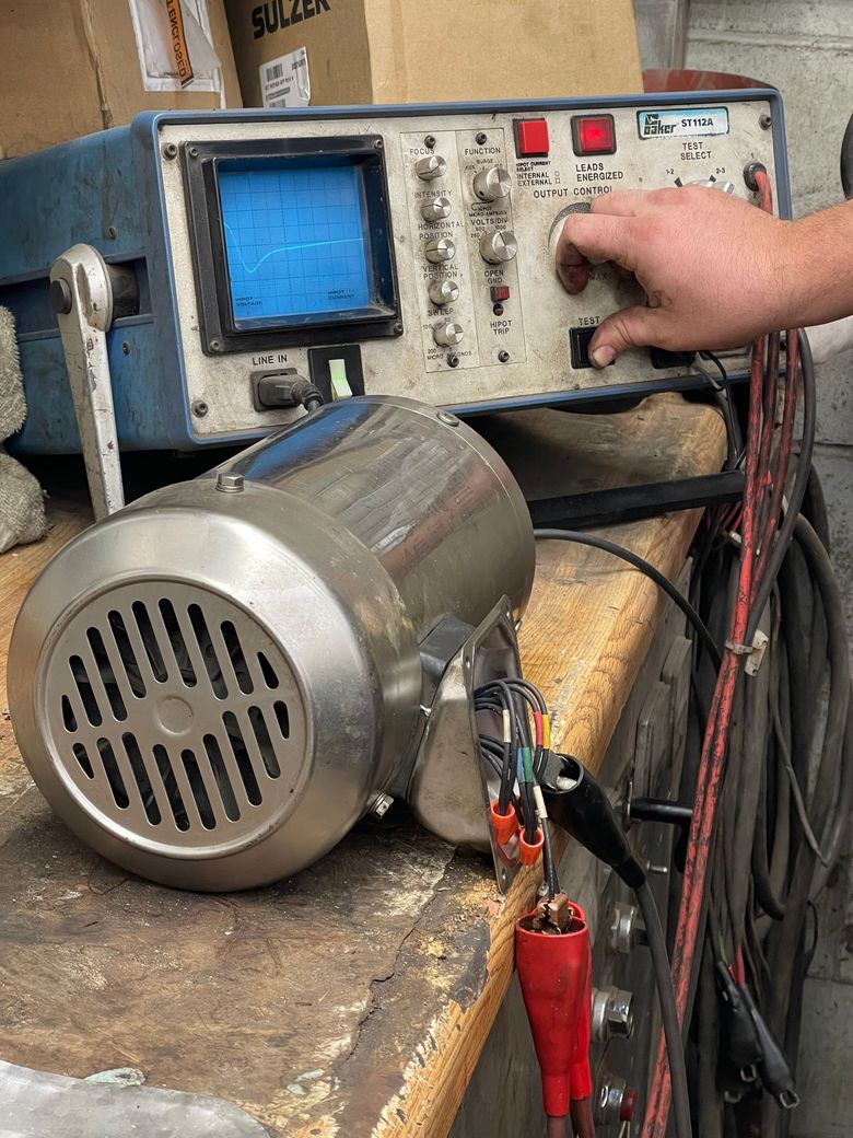A hand adjusts the dial on an oscilloscope connected to a cylindrical metal electric motor via jumper cables.