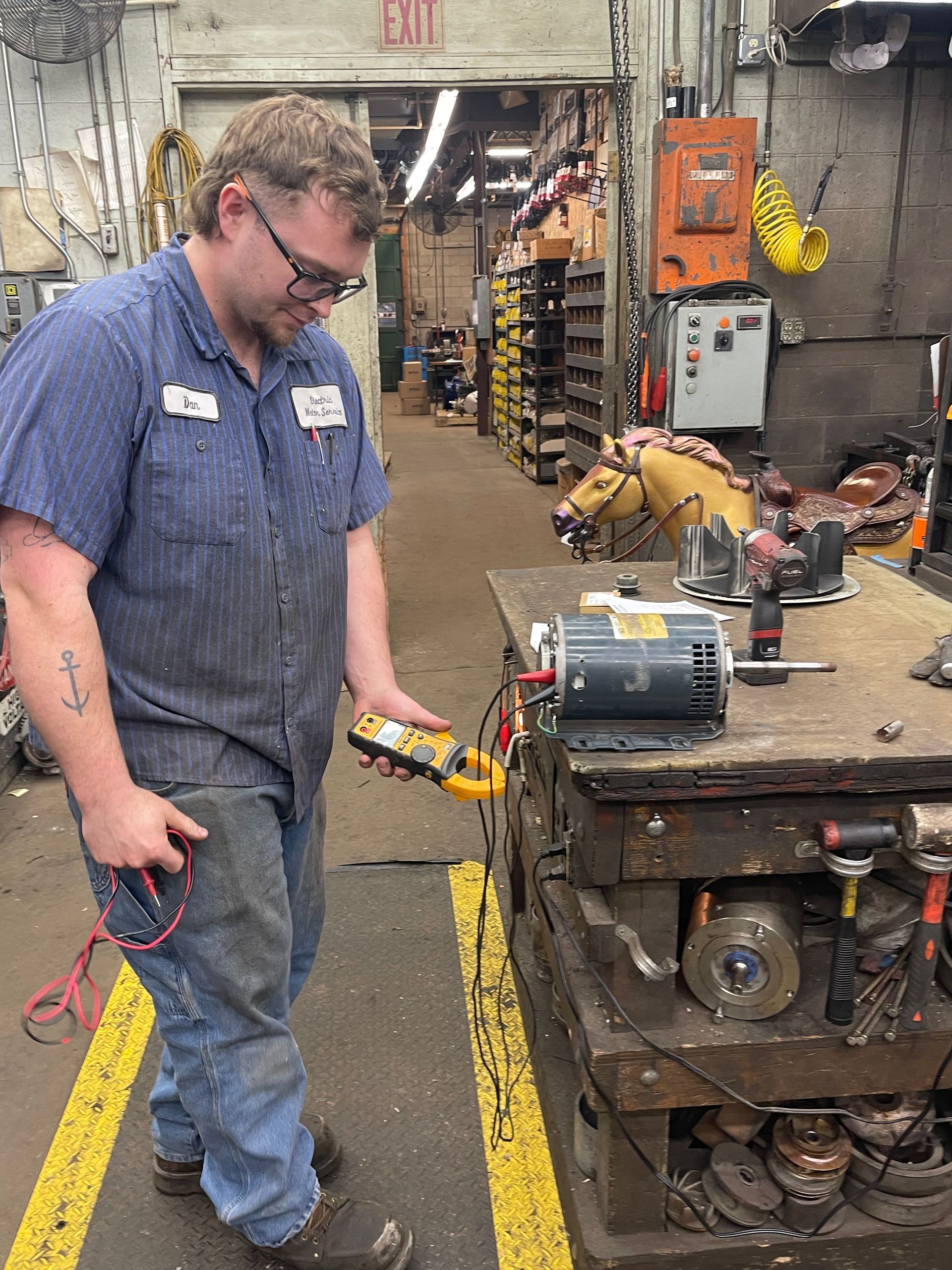 A worker in a blue uniform tests an electric motor on a workbench in a cluttered repair shop.