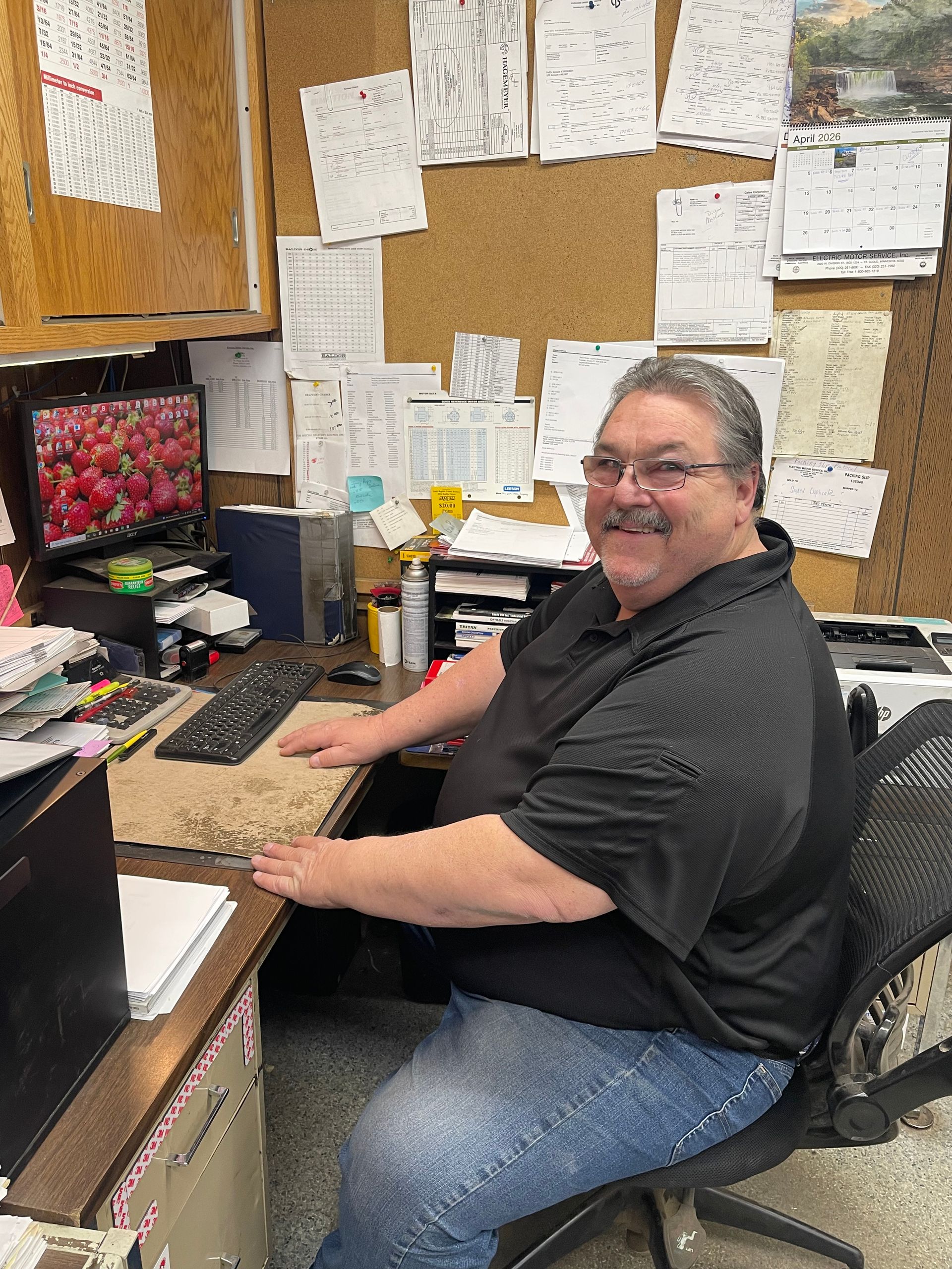 A smiling individual in a black polo shirt sits at a desk in an office, facing the camera with hands resting on a keyboard.