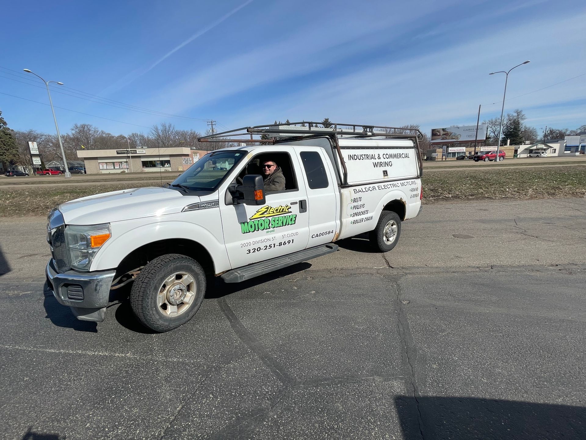 A white work truck with a ladder rack and company branding, parked on an asphalt lot under a clear blue sky.