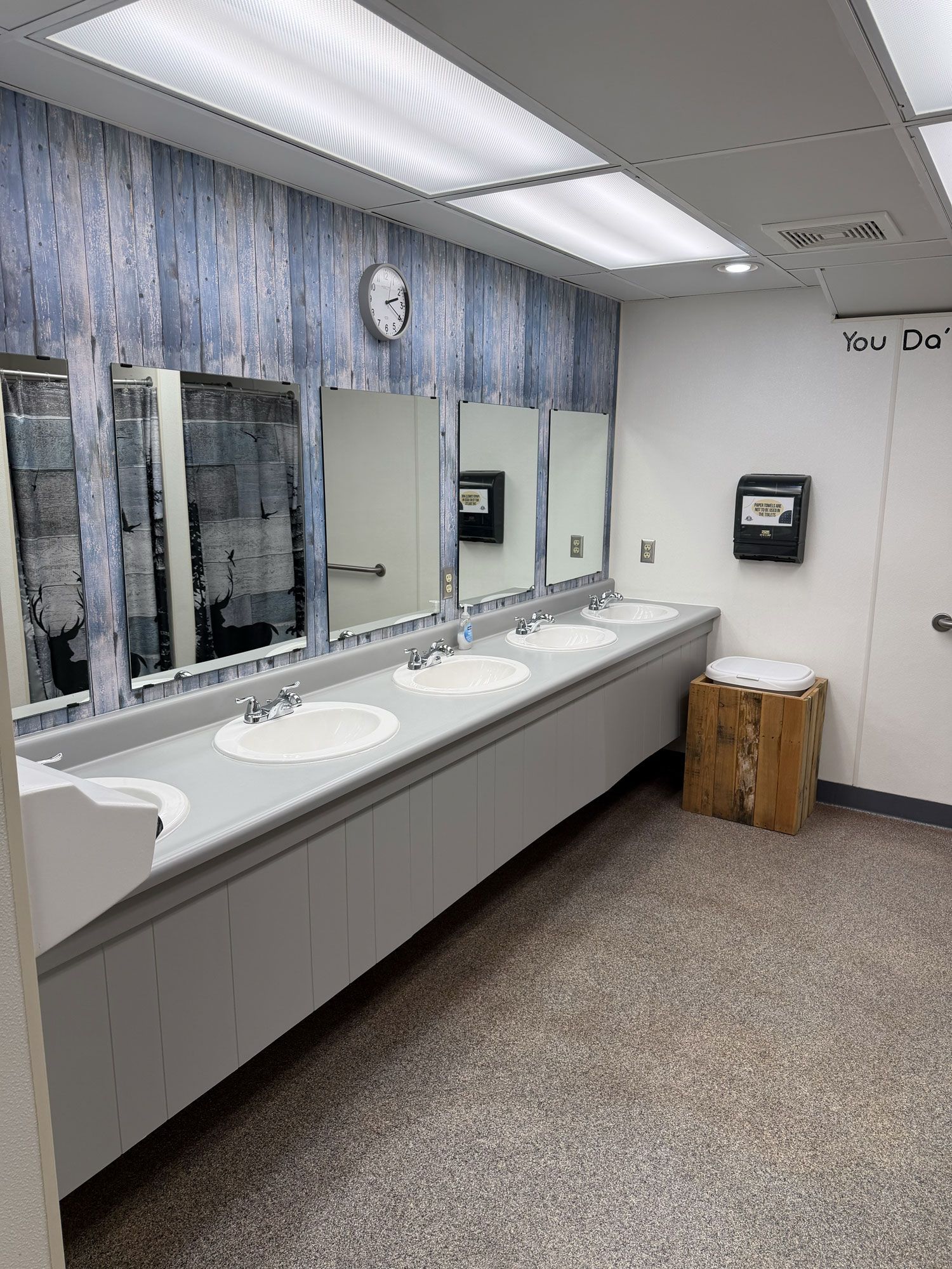 Public restroom with multiple sinks, mirrors, and wood-grain accent wall.
