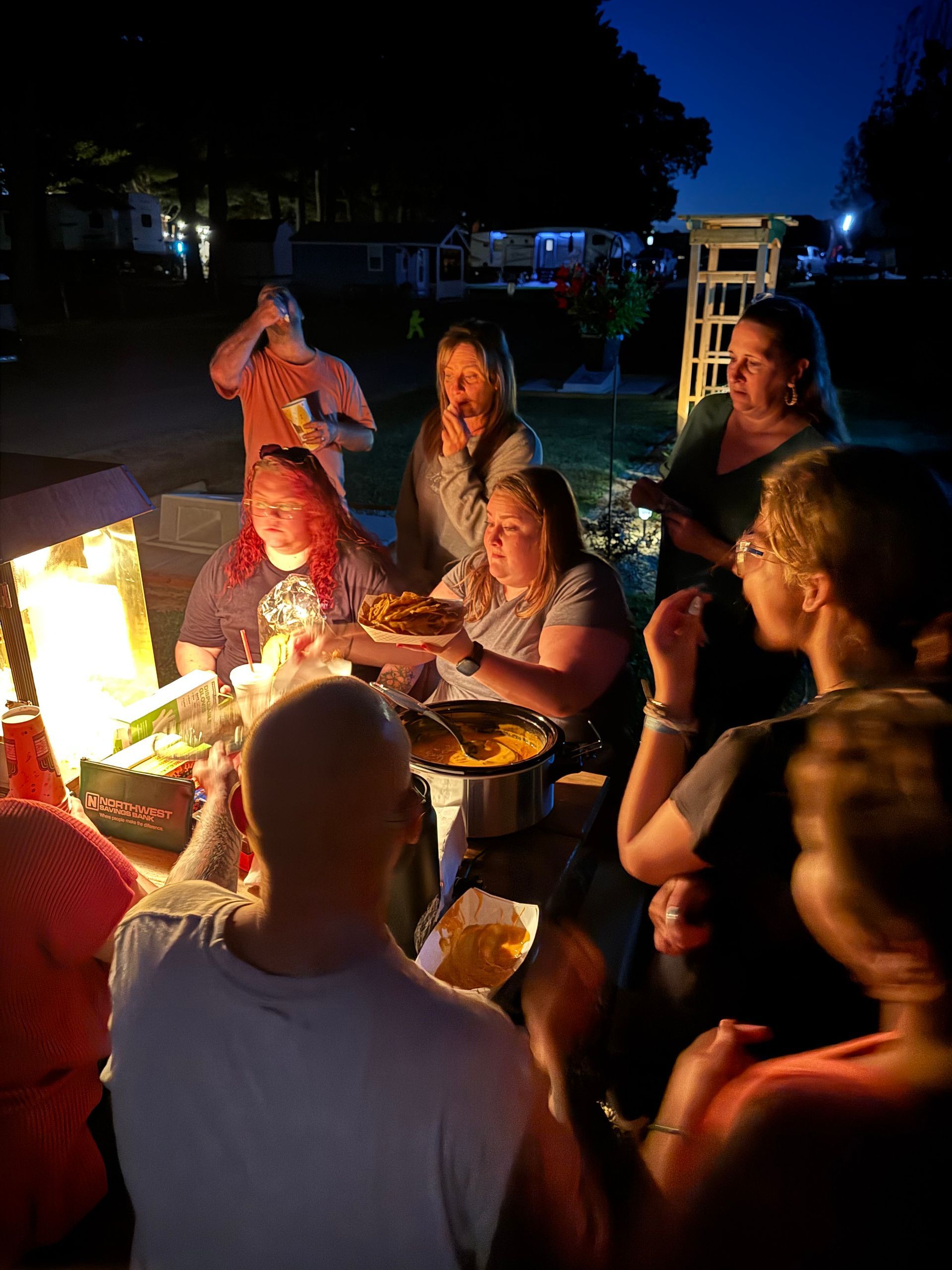 Group of people gathered around a table with food, lit by a grill and night lights, enjoying a meal outdoors.