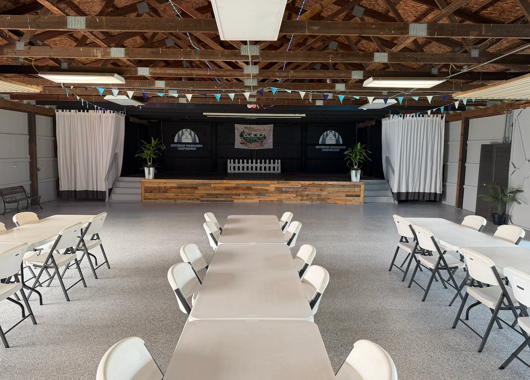 Long tables covered in blue and green cloths set up under a covered outdoor space; a speaker sits on a stage.