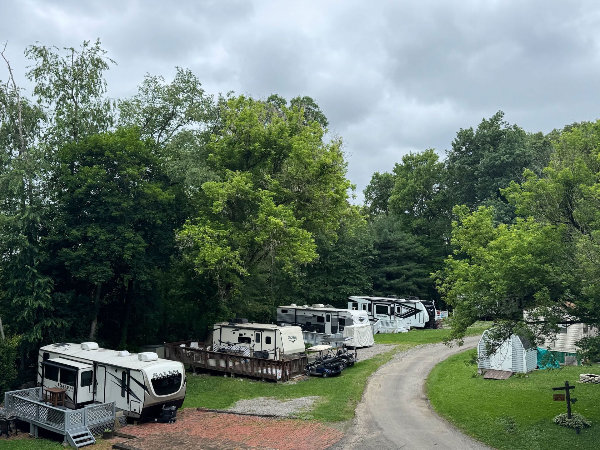 RVs parked in a campground surrounded by trees on a cloudy day; gravel road leads past them.