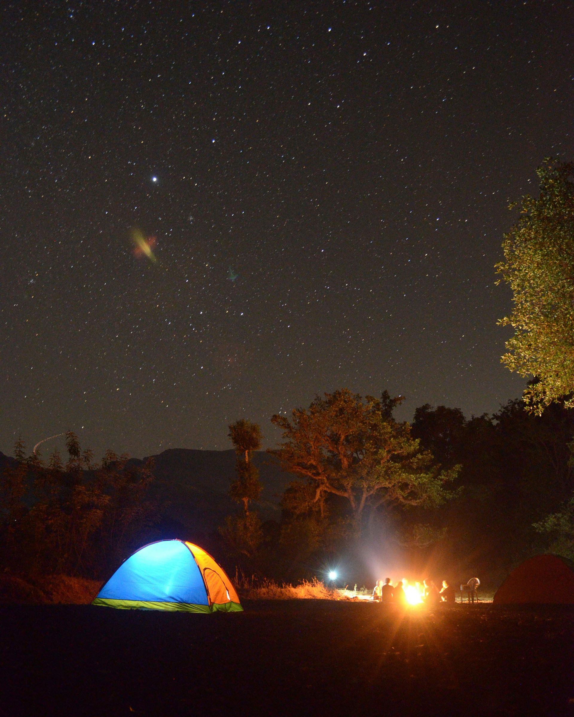Camping under starry night sky; tent glows blue, campfire burns orange, trees in the background.