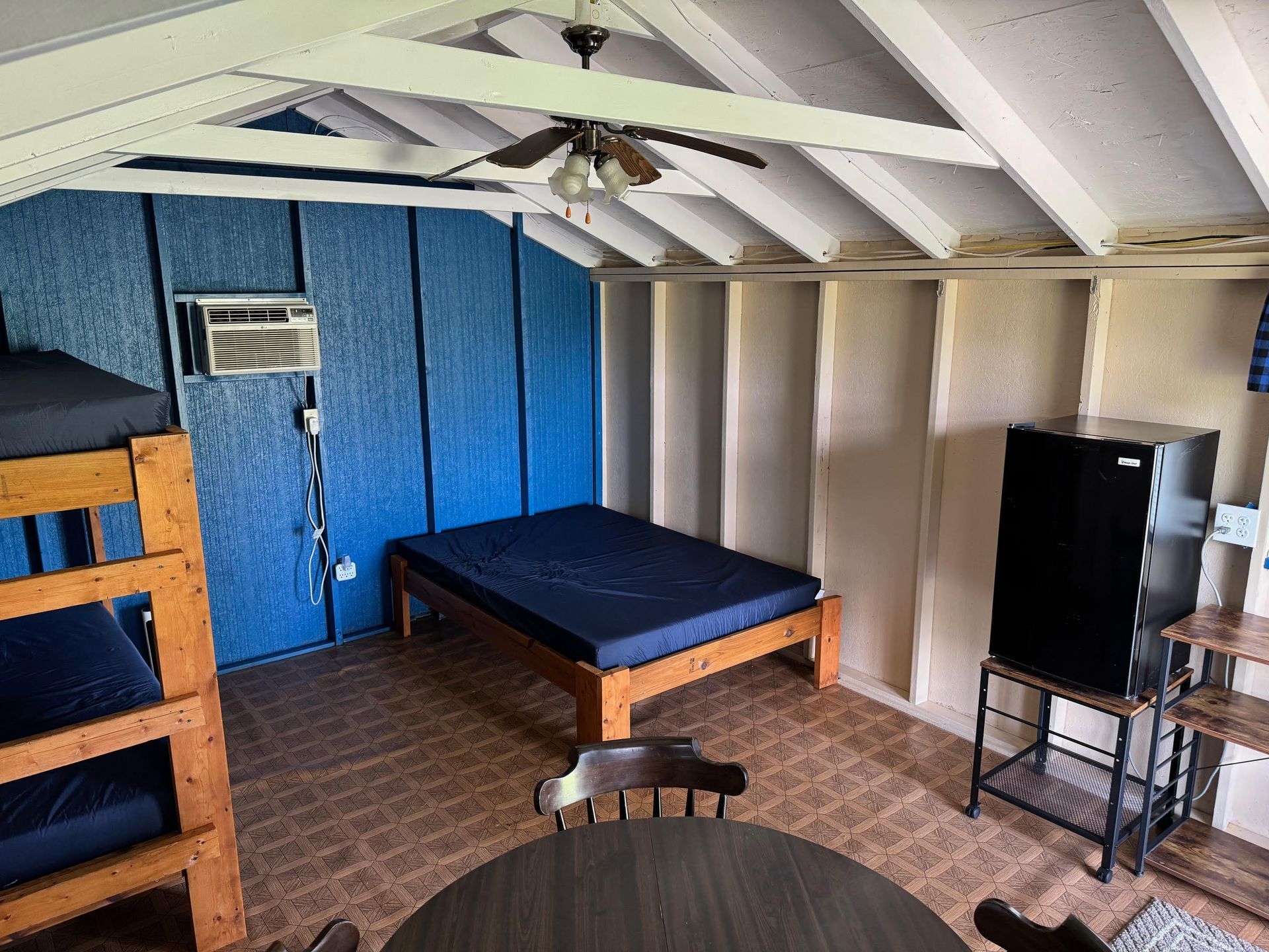 Interior of a small cabin with blue and white walls, bunk beds, a bed, a small refrigerator, and a ceiling fan.