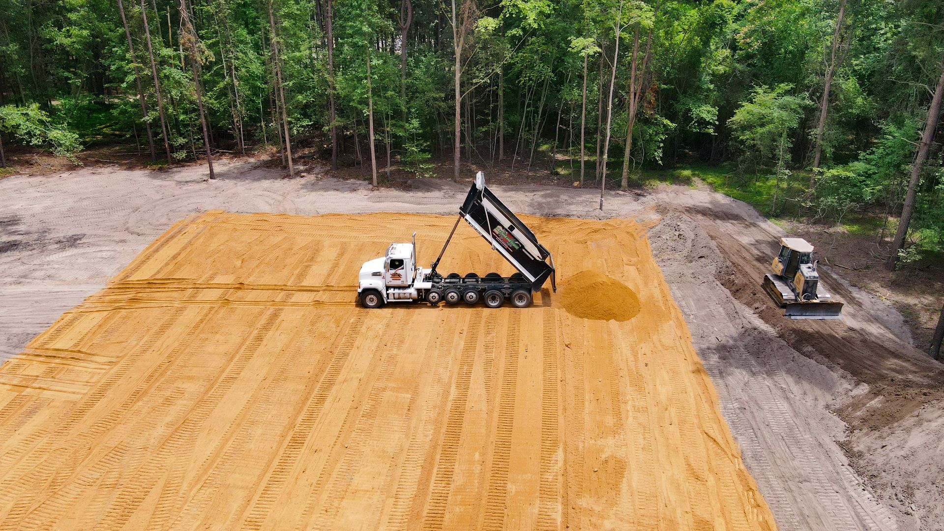 Dump truck unloading dirt onto a graded construction site in a wooded area.
