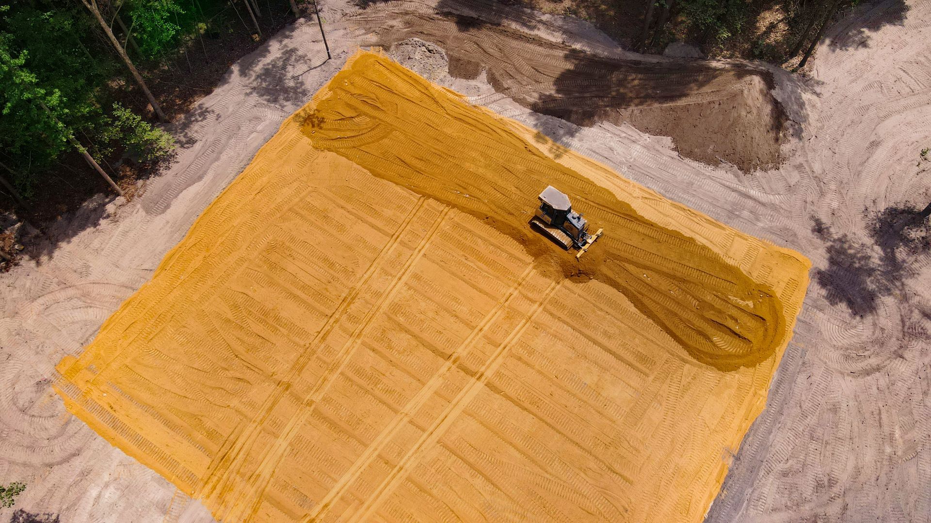 Aerial view of a bulldozer leveling soil in a large, sandy clearing near trees.