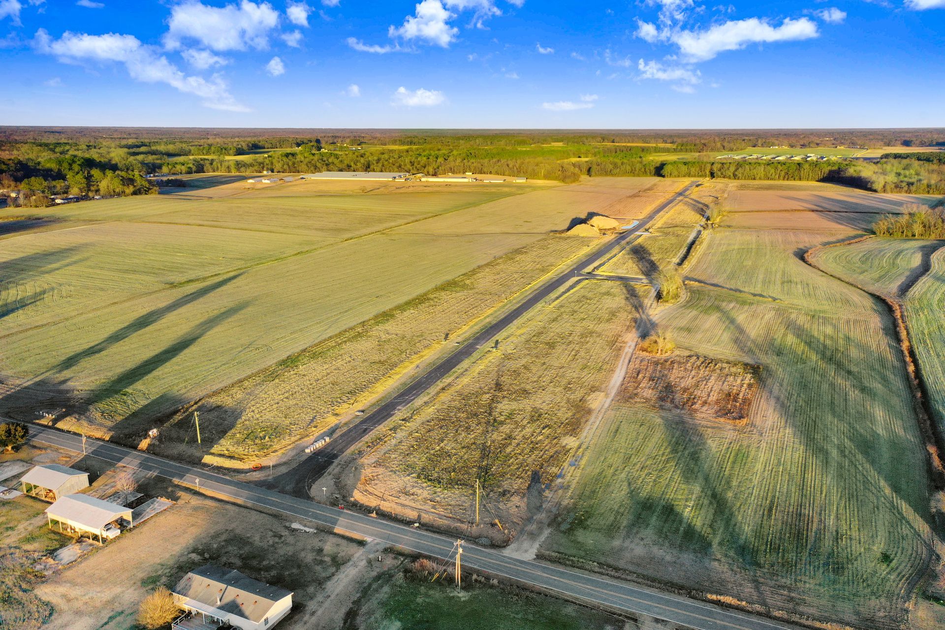 Aerial view of a rural landscape with fields, a road, and buildings under a blue sky.