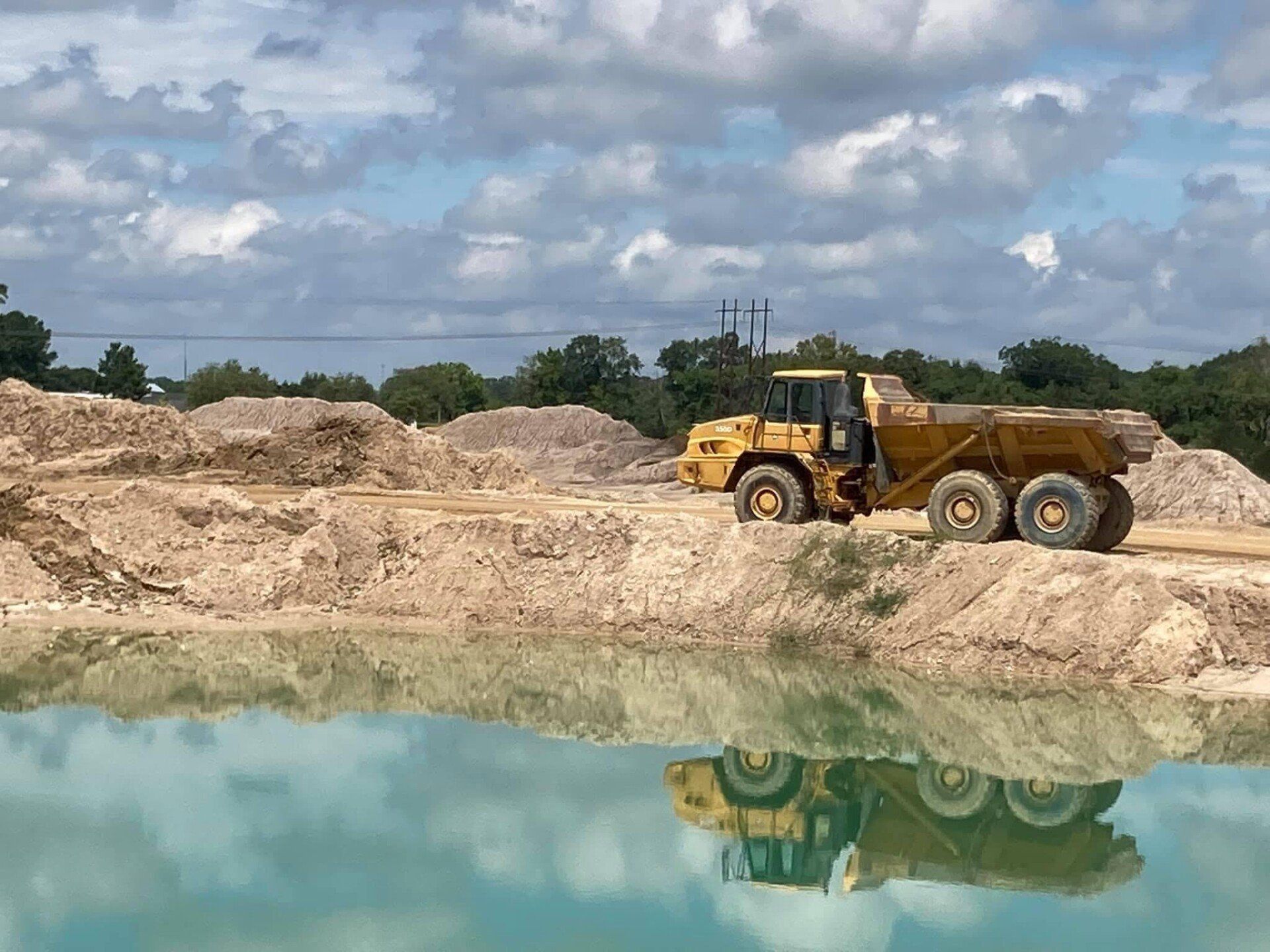 Dump truck at the edge of a water feature, reflecting its yellow color. Cloudy sky in background.