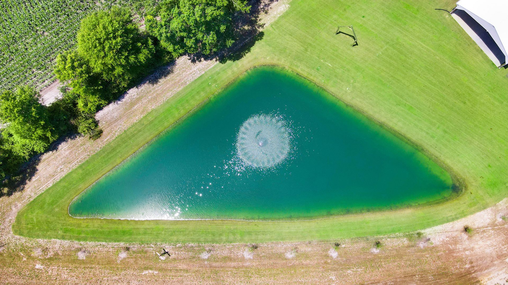Aerial view of a triangular pond with aerator, surrounded by green grass and a field.