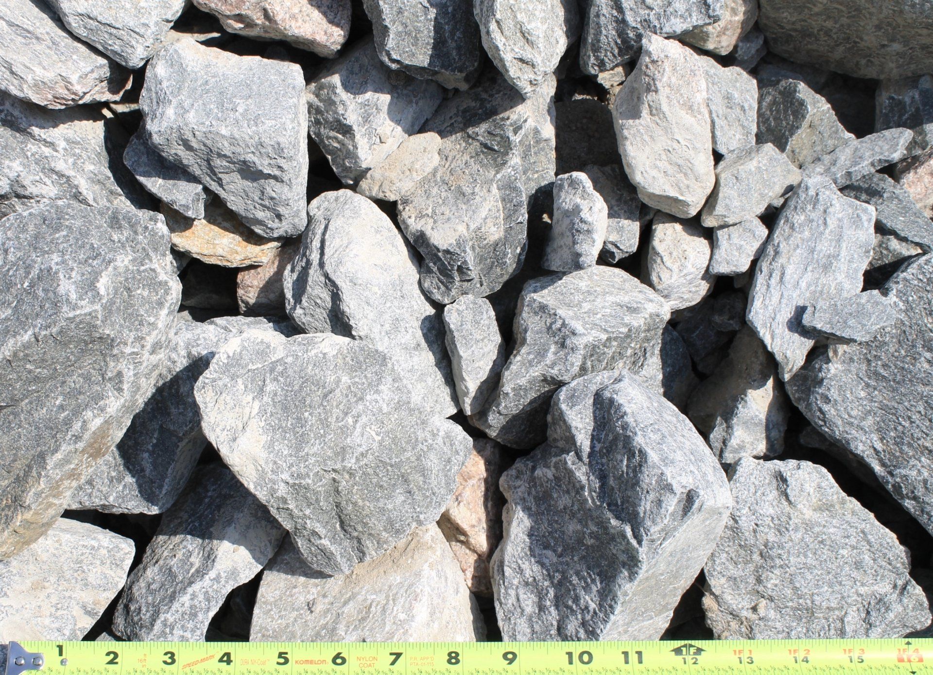 Pile of gray, angular rocks of various sizes, with a yellow and green ruler at the bottom.