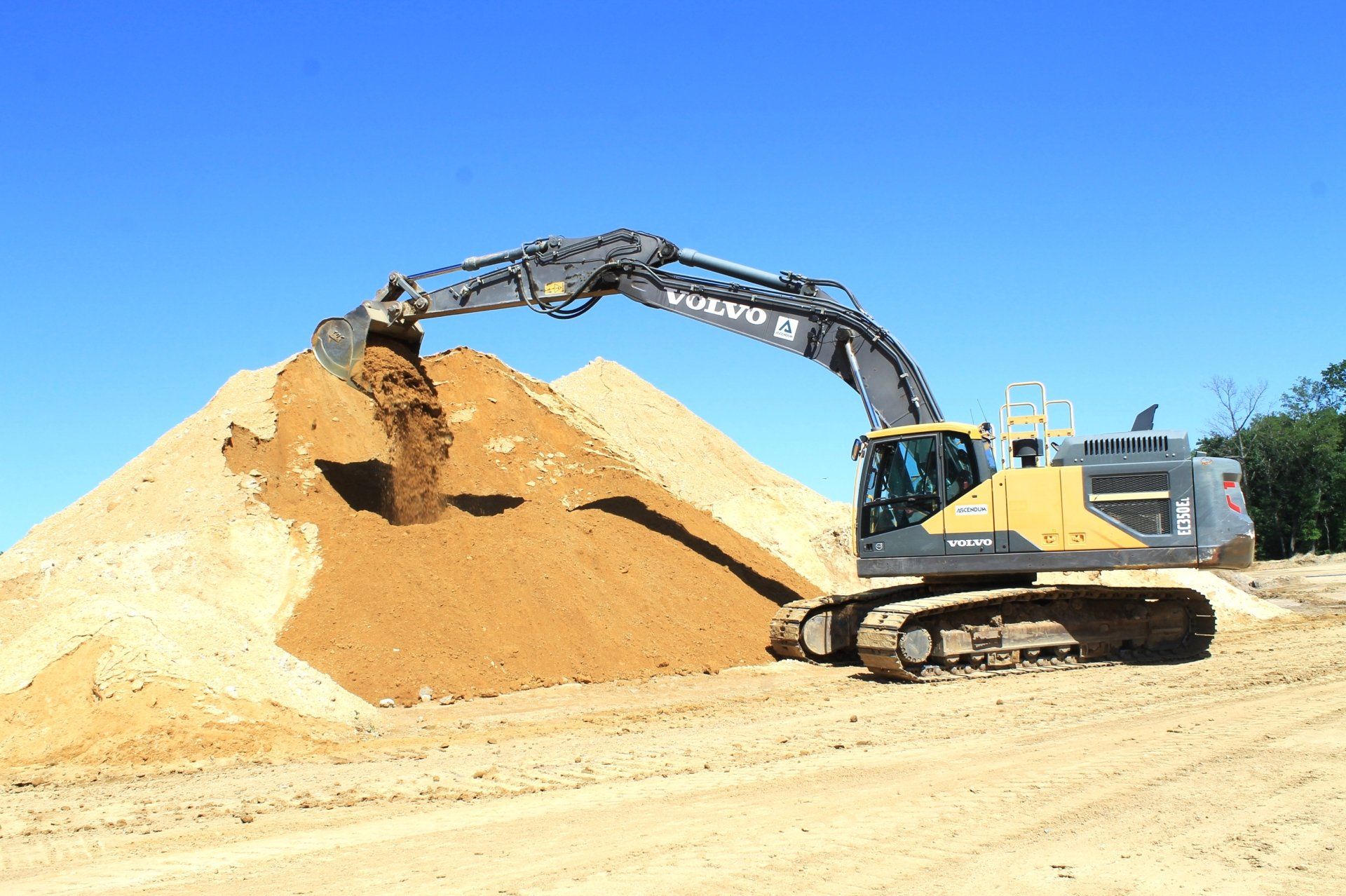 An excavator dumping soil onto a large pile outdoors under a blue sky.