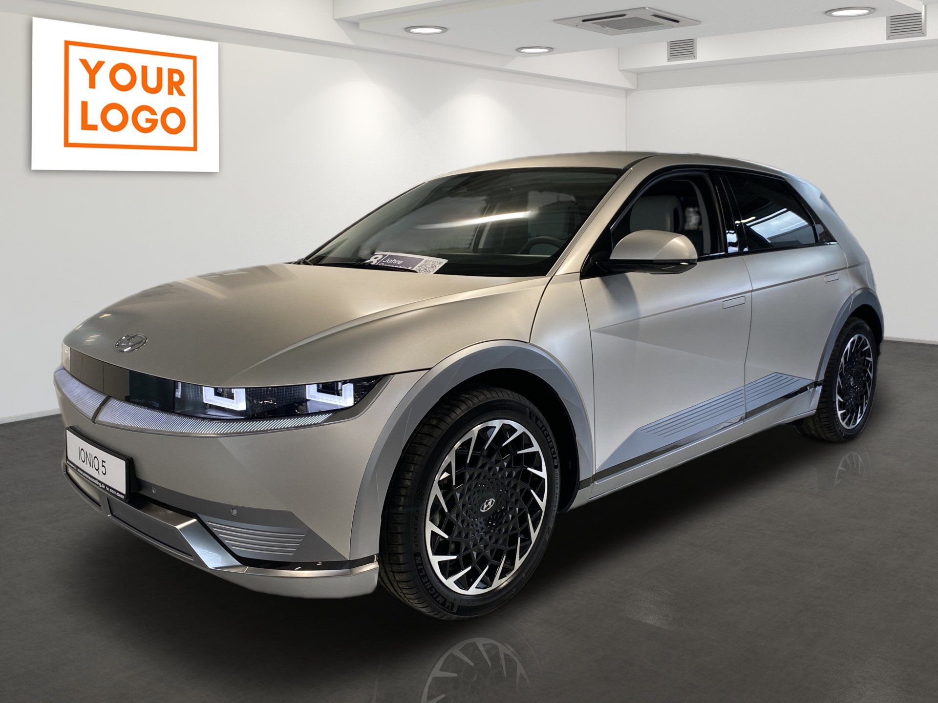 Silver electric hatchback in a showroom with a “Your Logo” sign on the wall