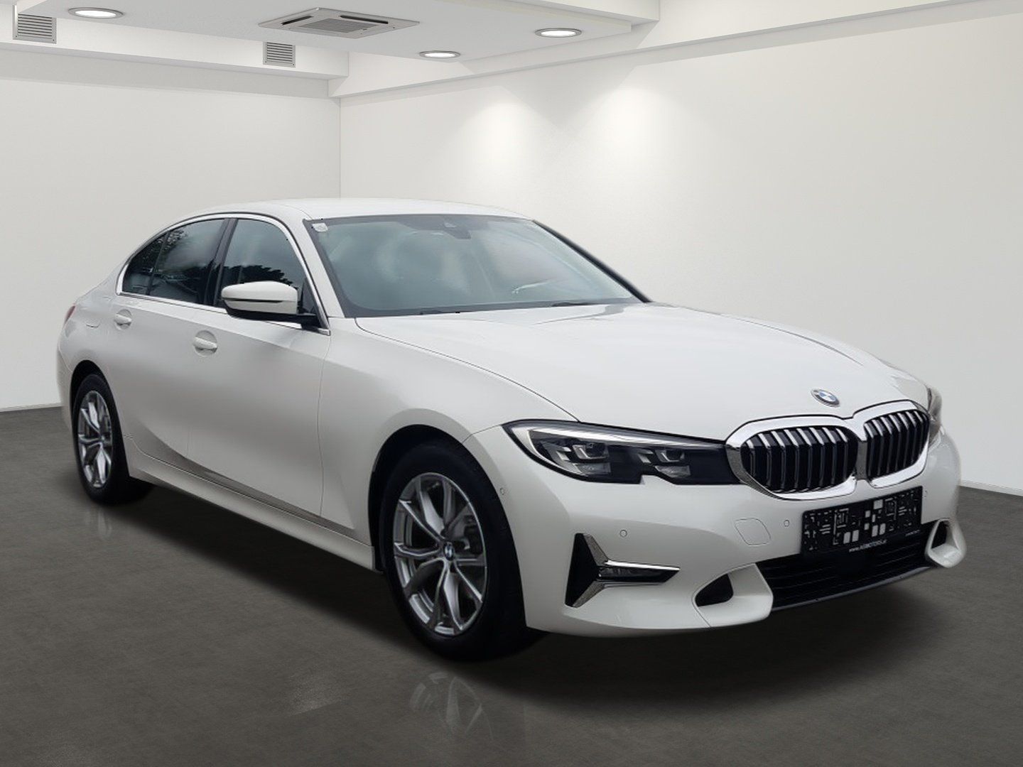 Biely sedan BMW zaparkovaný v presvetlenom štúdiovom showroome, pohľad spredu z troch štvrtín.