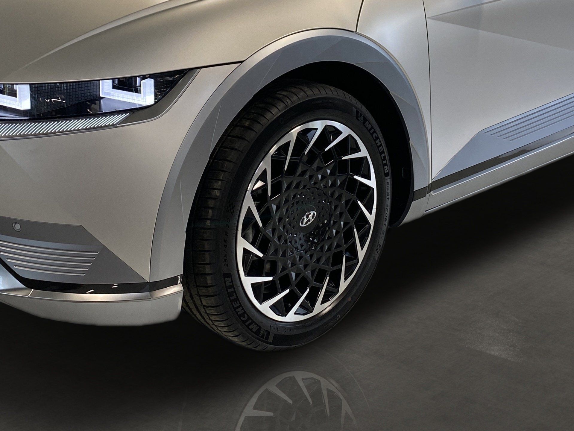 Every detail visible in your digital showroom Close-up of a silver car’s front wheel and bumper on a dark showroom floor