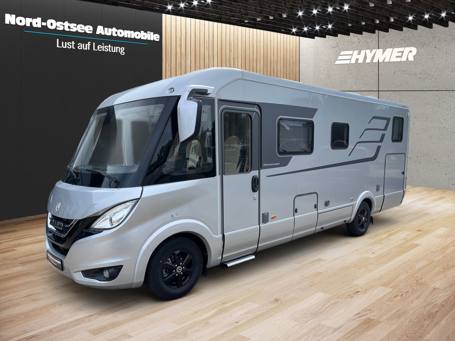 Strieborný obytný automobil vystavený v interiéri na autosalóne, pohľad z ľavej prednej strany.
