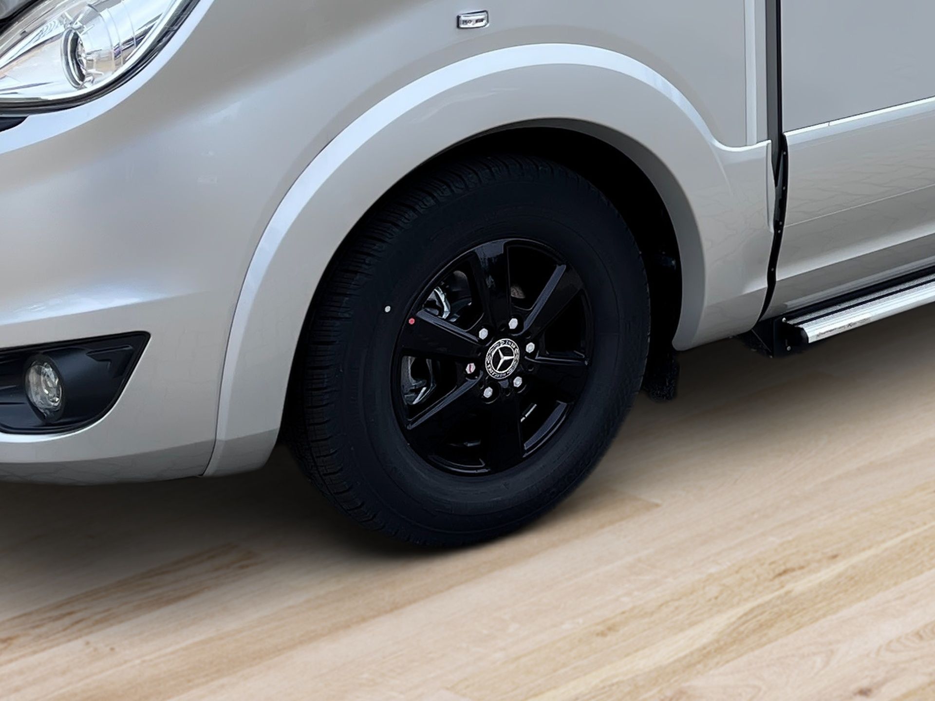 Detailný záber predného kolesa strieborného SUV s čiernym ráfikom na svetlej drevenej podlahe.