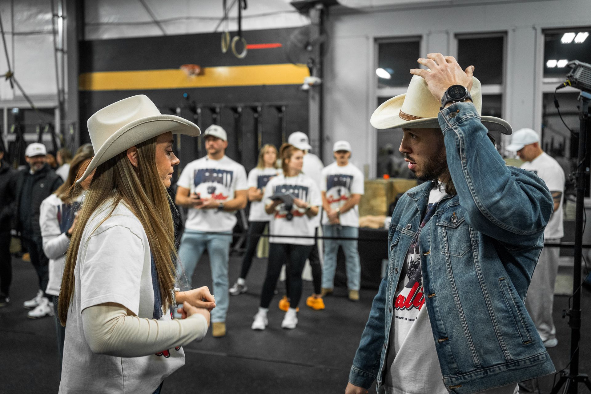 Two people in cowboy hats talking in a gym, with a line of people standing in the background.