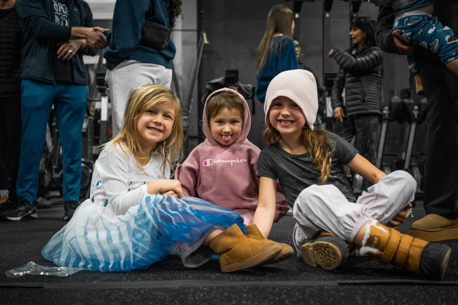 Three children sit smiling on a curb, two in costumes, with adults standing behind them.