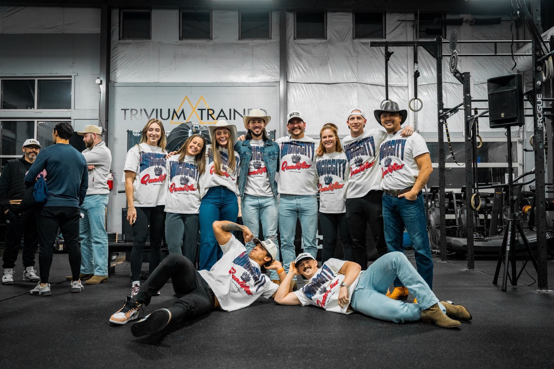 Group posing in matching white shirts inside a gym, with two people lying on the floor in front.