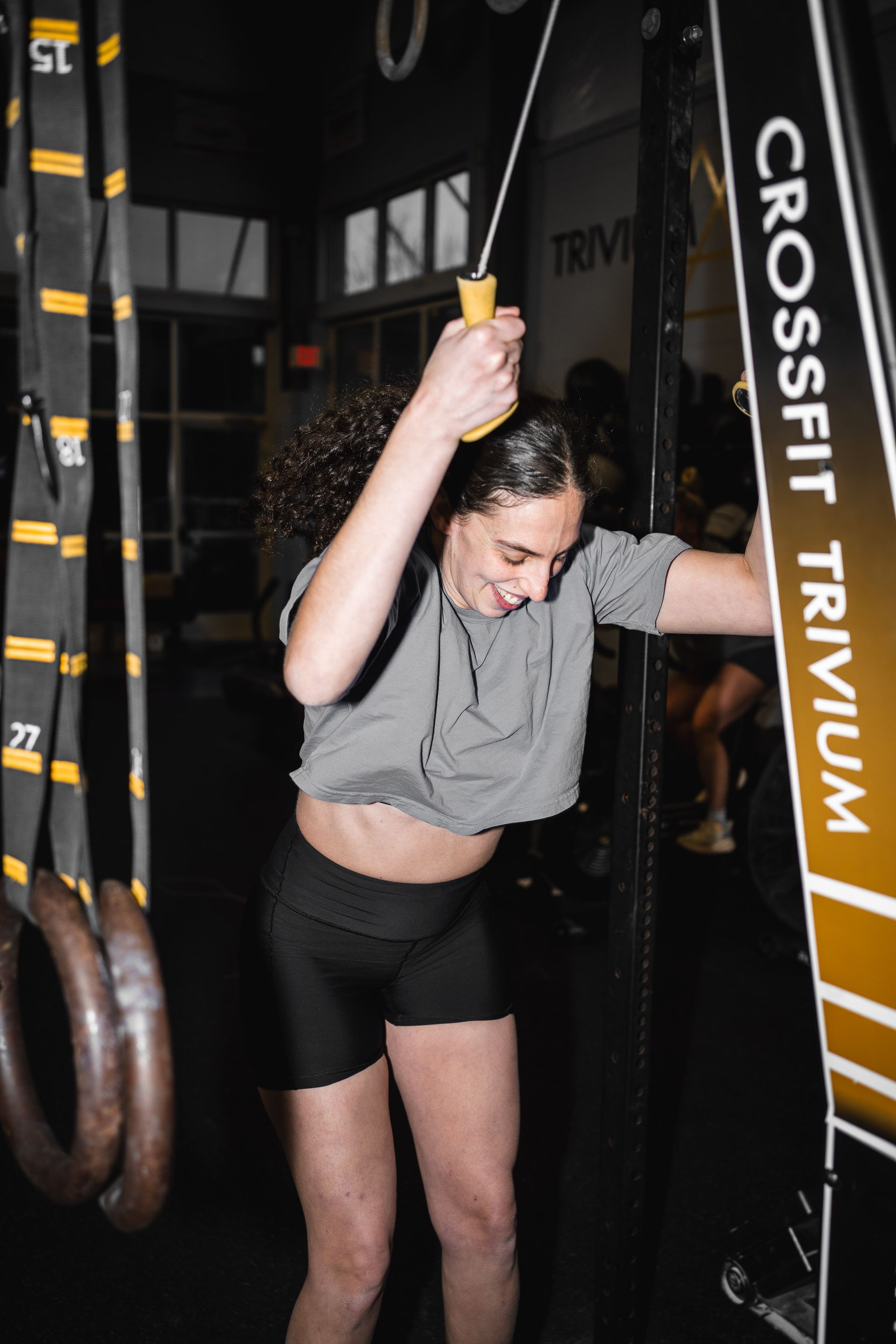 Person in a gym adjusting a hanging TRX strap, wearing a gray shirt and black shorts.