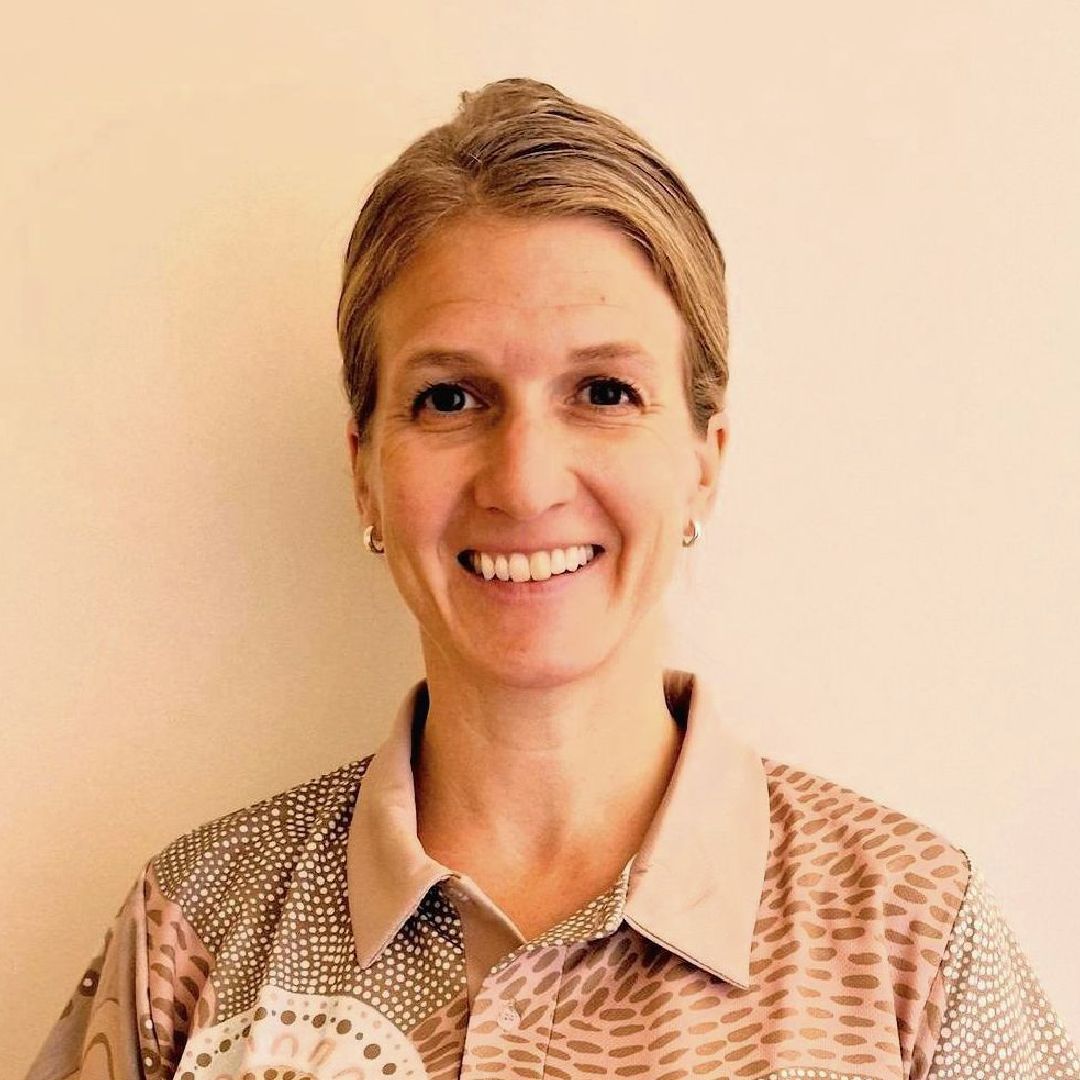 Smiling person with short blond hair wearing a patterned collared shirt against a beige background