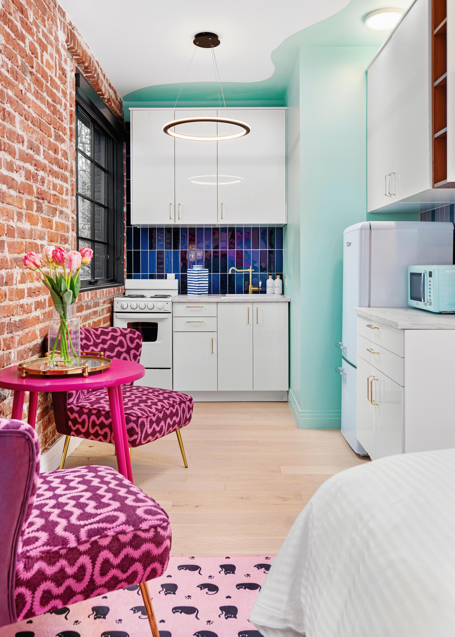 Bright compact kitchen with pink accents, brick wall, turquoise cabinet, and white appliances