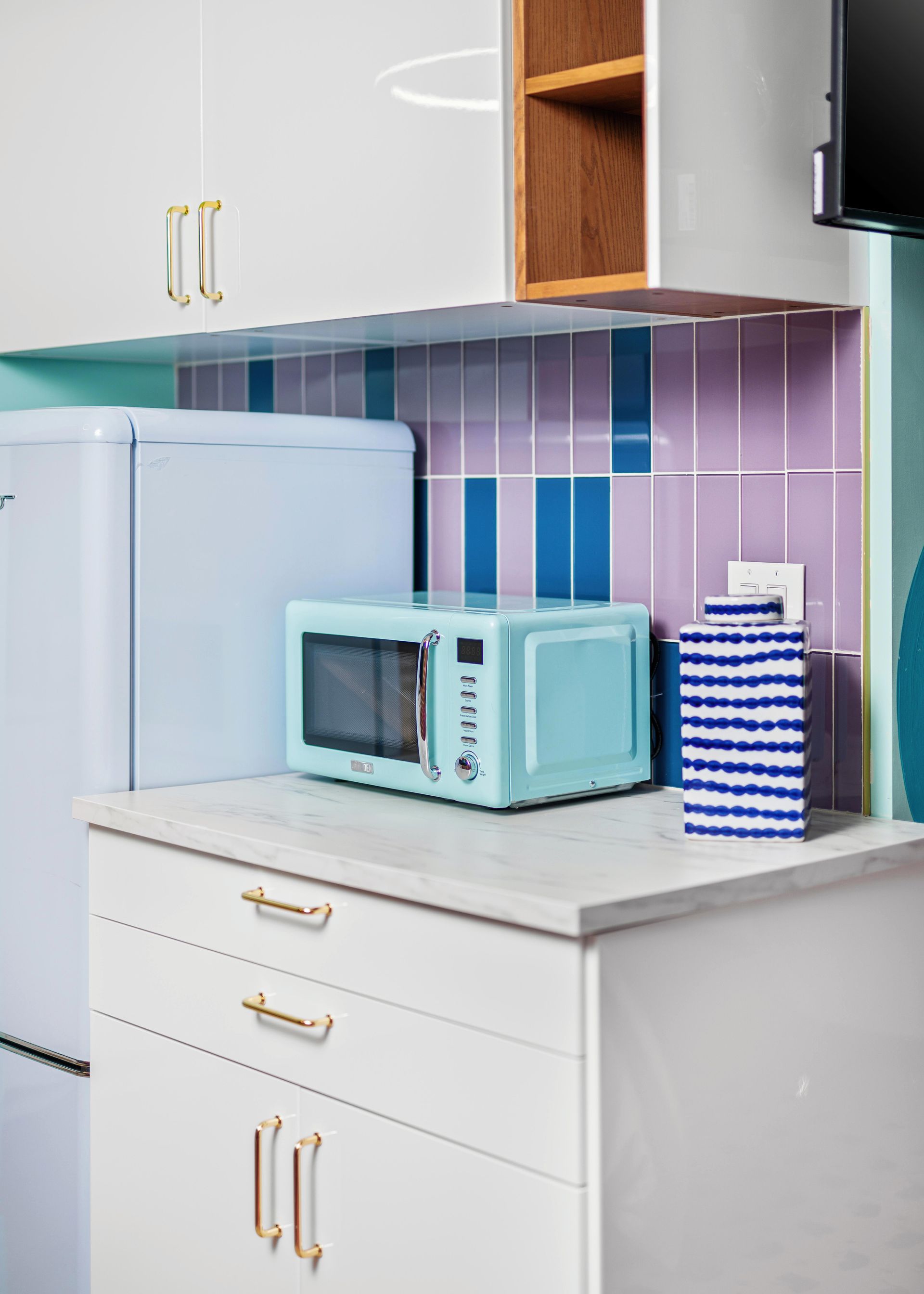 Compact kitchen with pastel blue microwave on a white counter, beside a fridge and striped backsplash.
