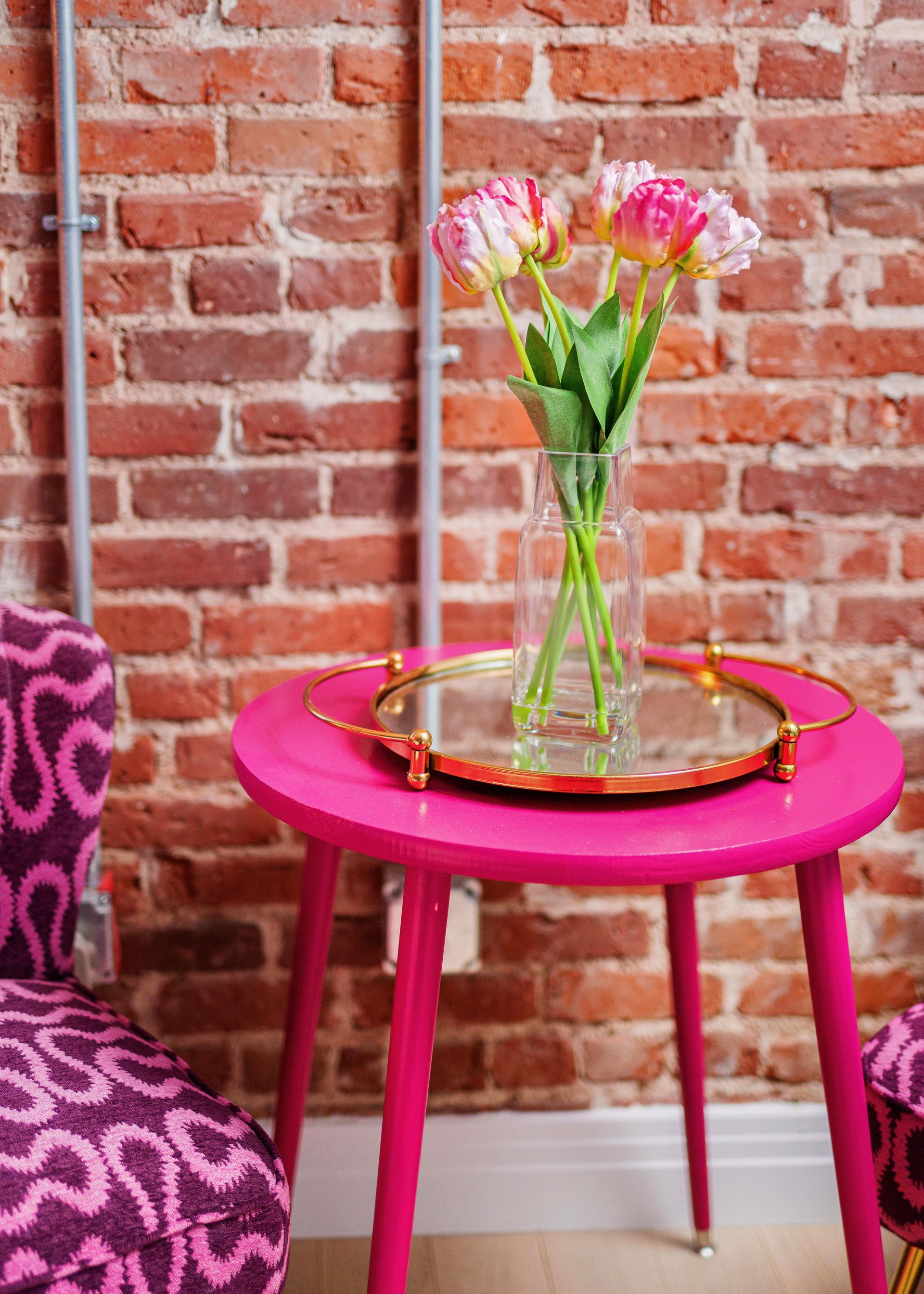 Pink side table with a glass vase of flowers and a gold tray against a brick wall.