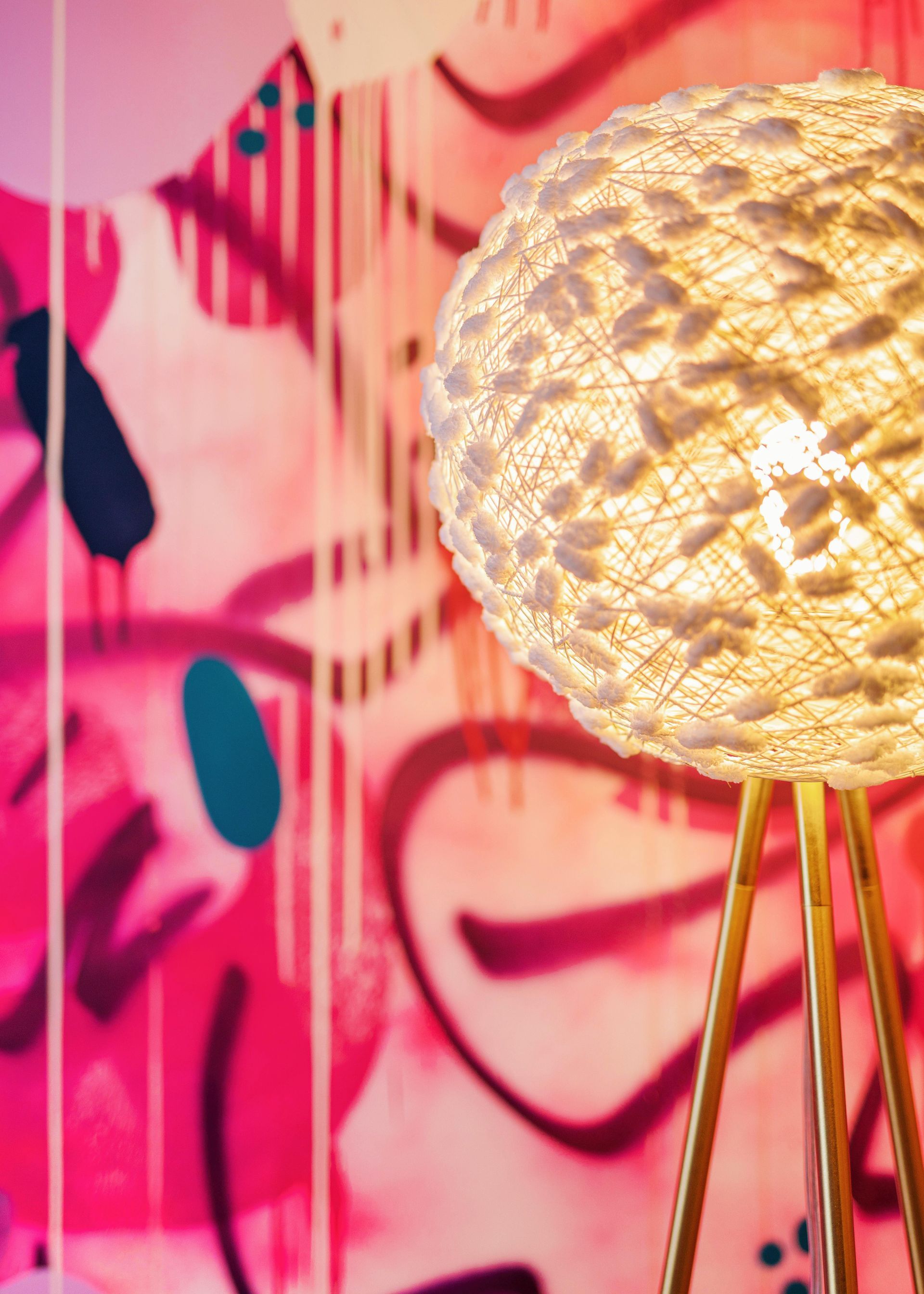 Woven globe floor lamp glowing beside a pink wall with abstract art and face-like graffiti