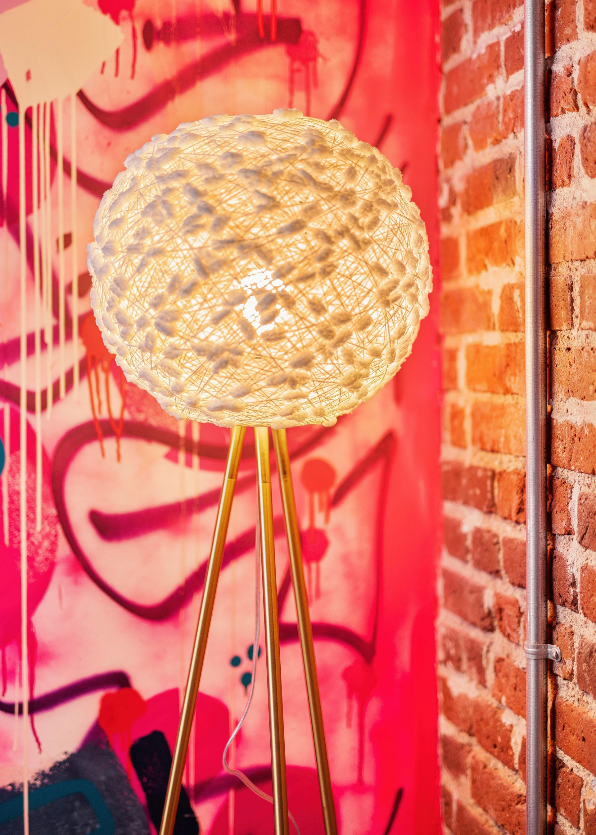 Large woven sphere lamp on a thin stand against a pink graffiti wall and brick backdrop.