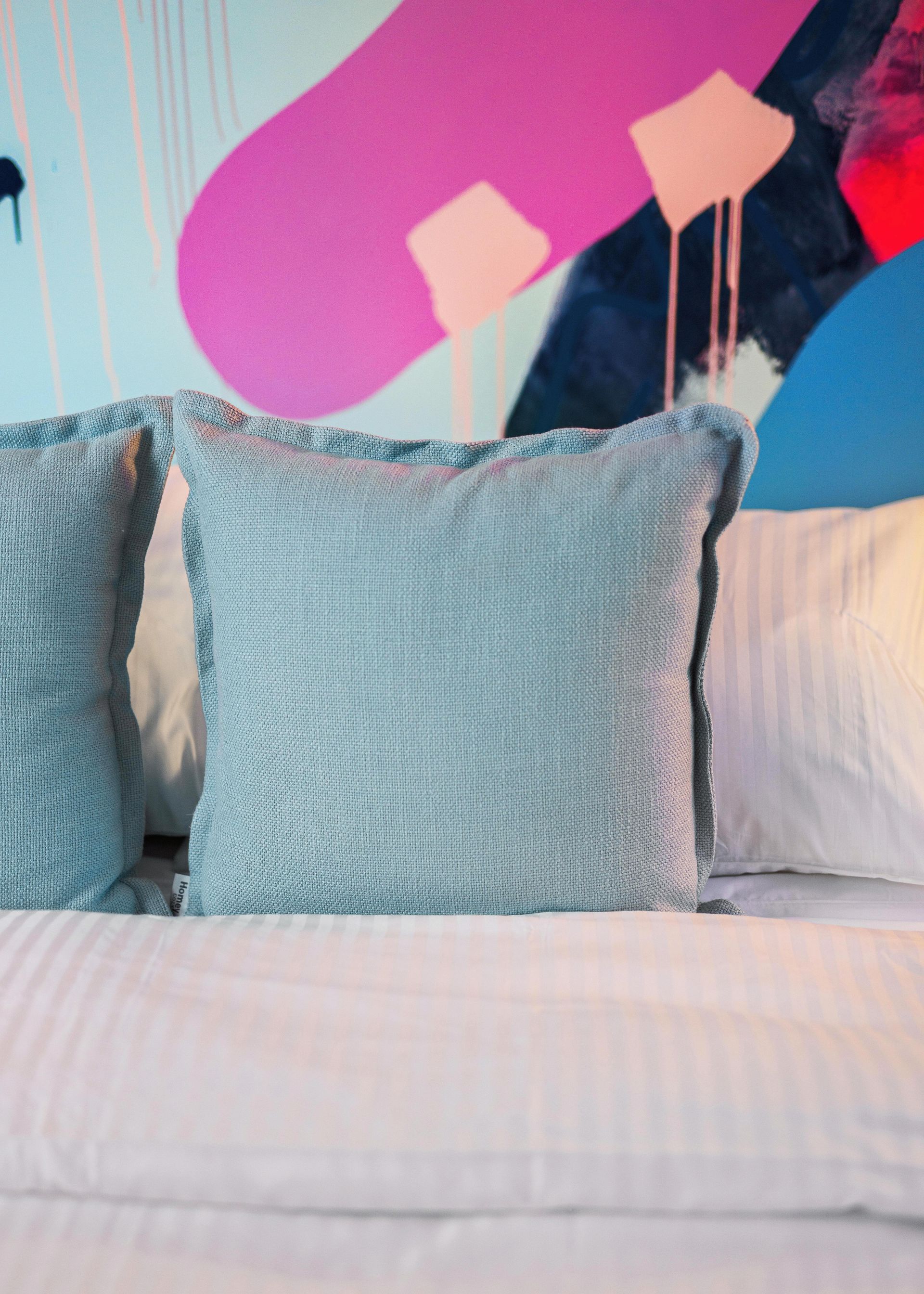 Blue pillows on a white bed against a colorful abstract wall mural.