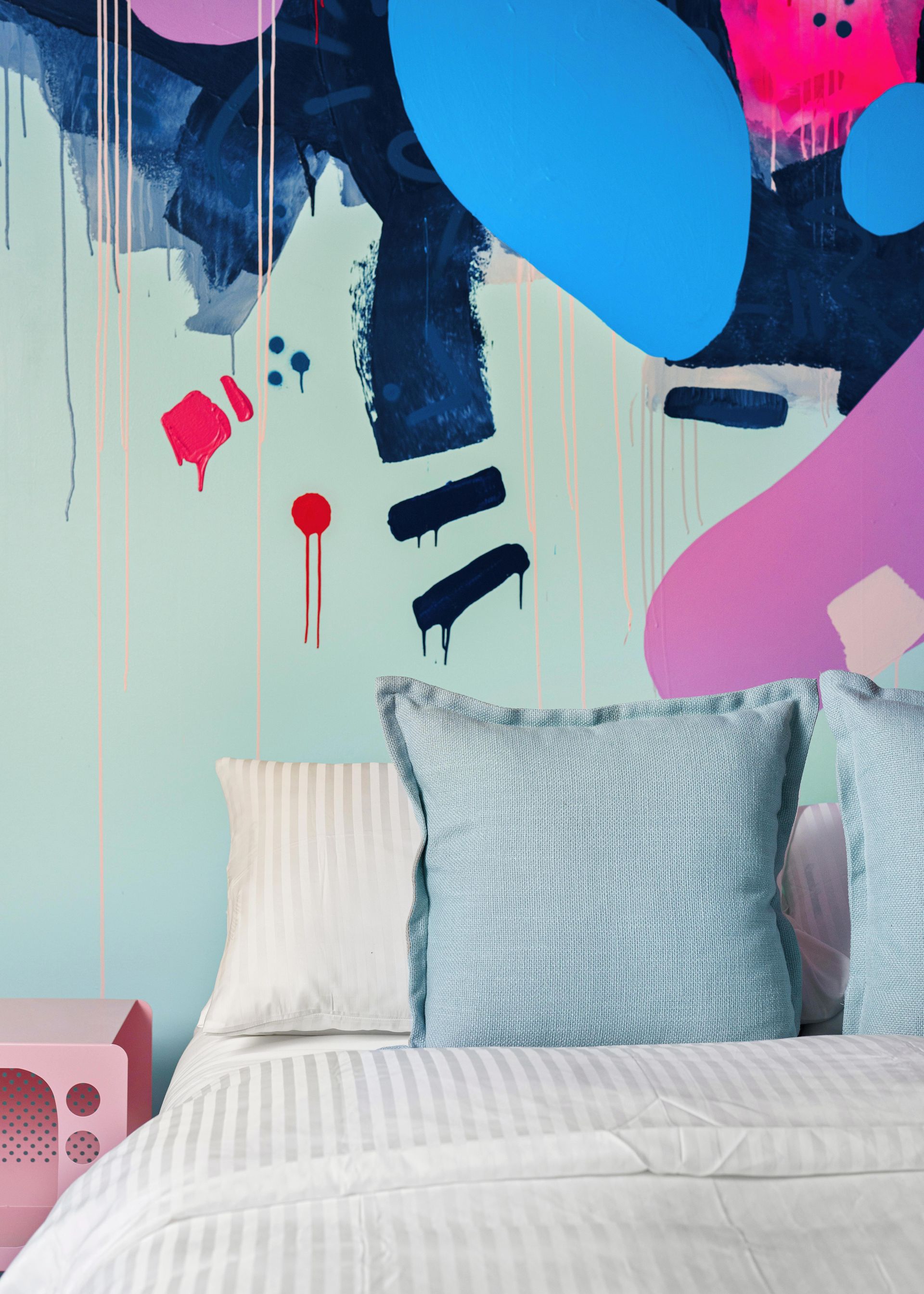 Colorful abstract mural behind a neatly made bed with white bedding and a blue textured pillow