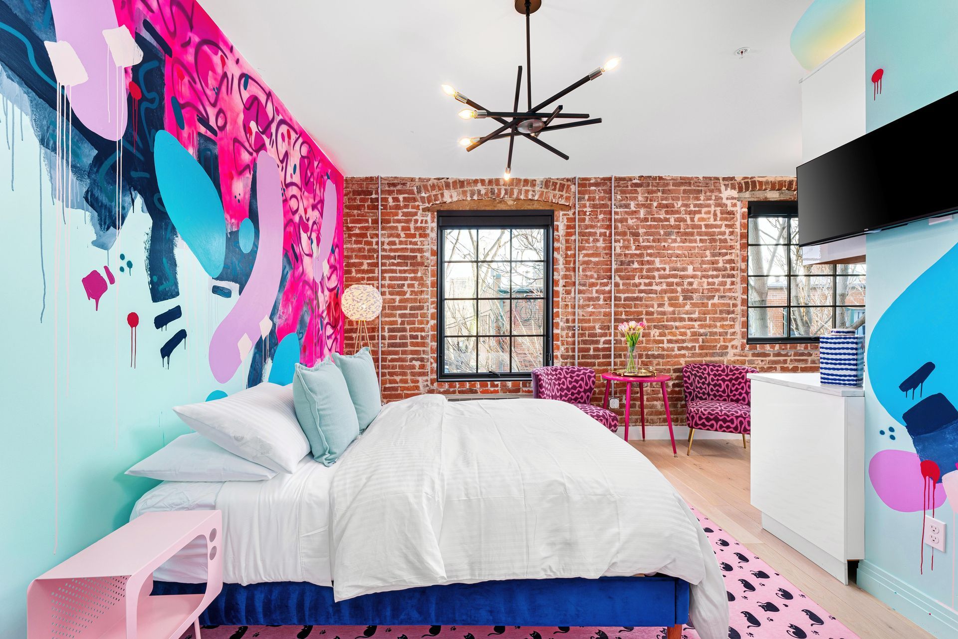 Bright bedroom with colorful wall mural, white bed, exposed brick wall, and large window.