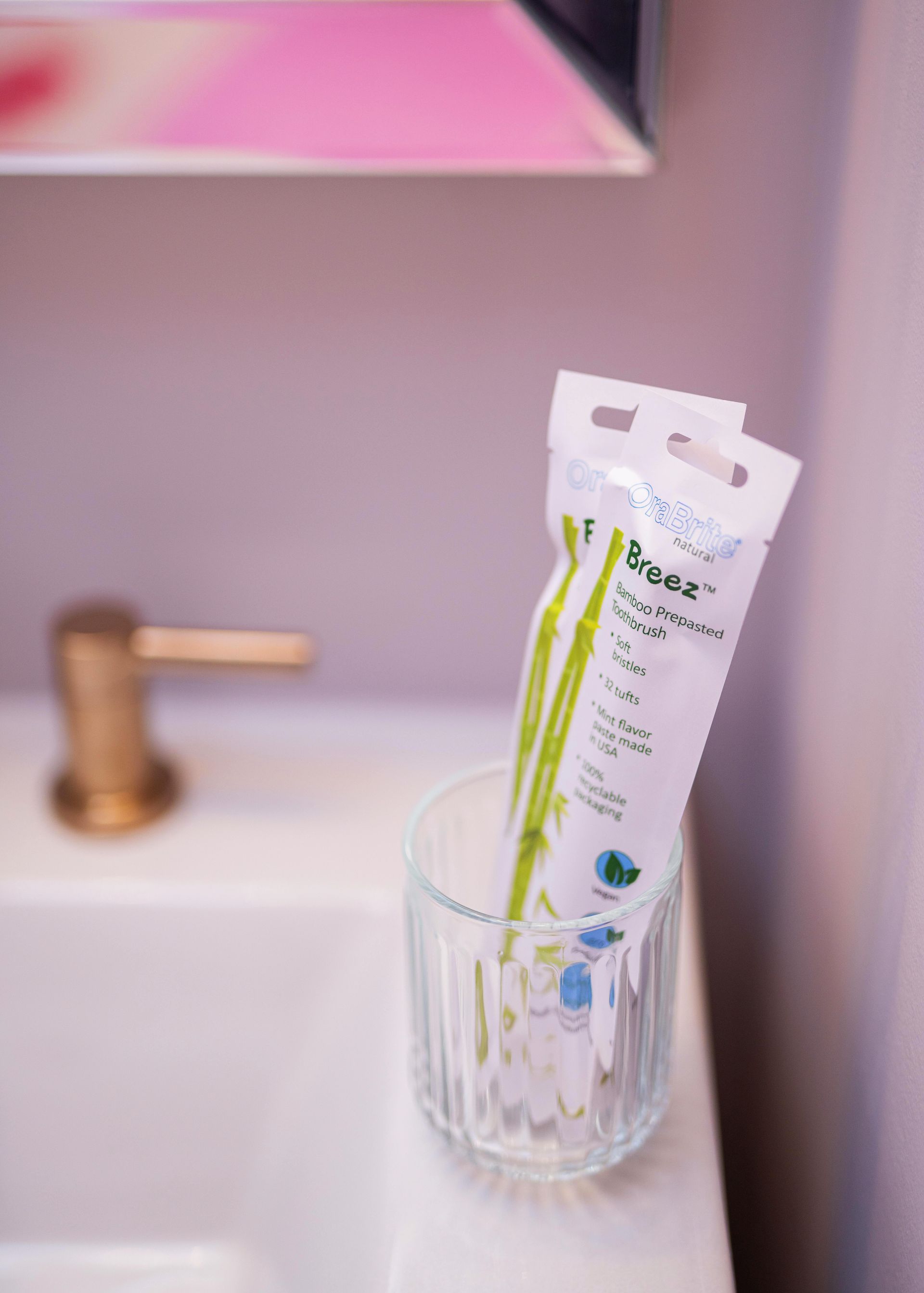 Toothbrushes in a clear glass beside a pink sink and gold faucet