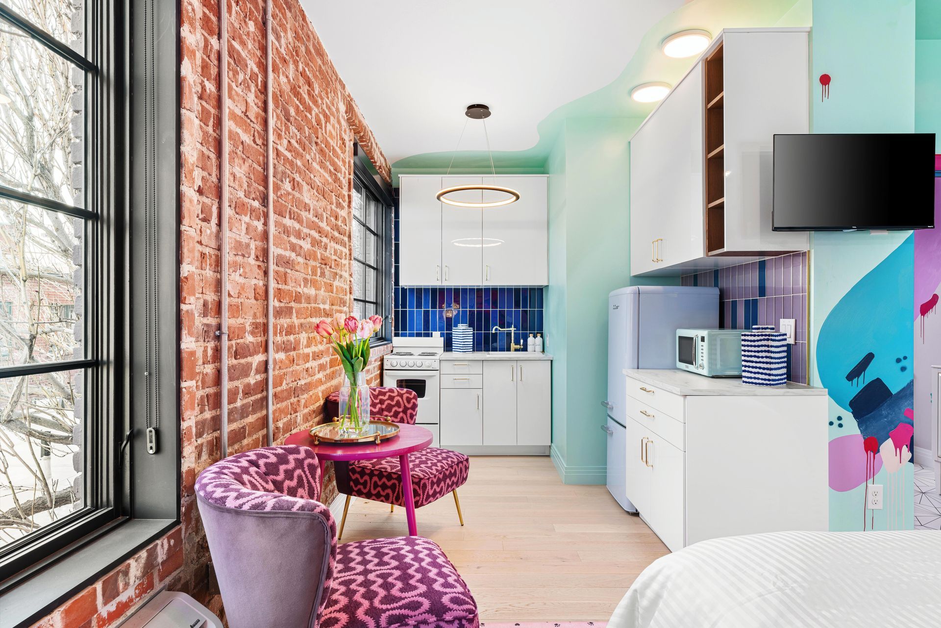Bright narrow living room with brick wall, colorful furniture, kitchenette, and wall-mounted TV.