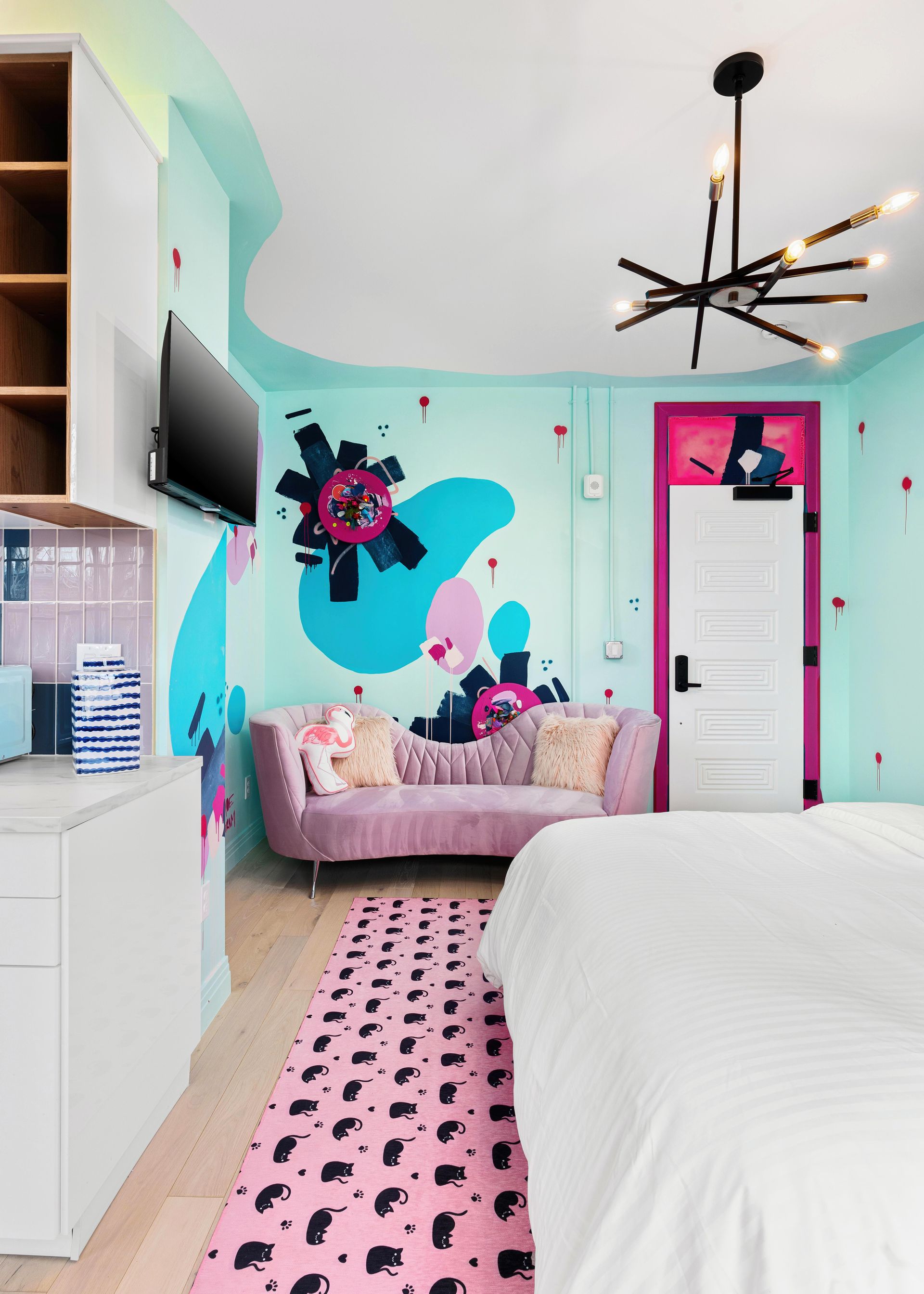 Pastel pink and teal bedroom with a bed, sofa, floral wall art, and modern black chandelier.