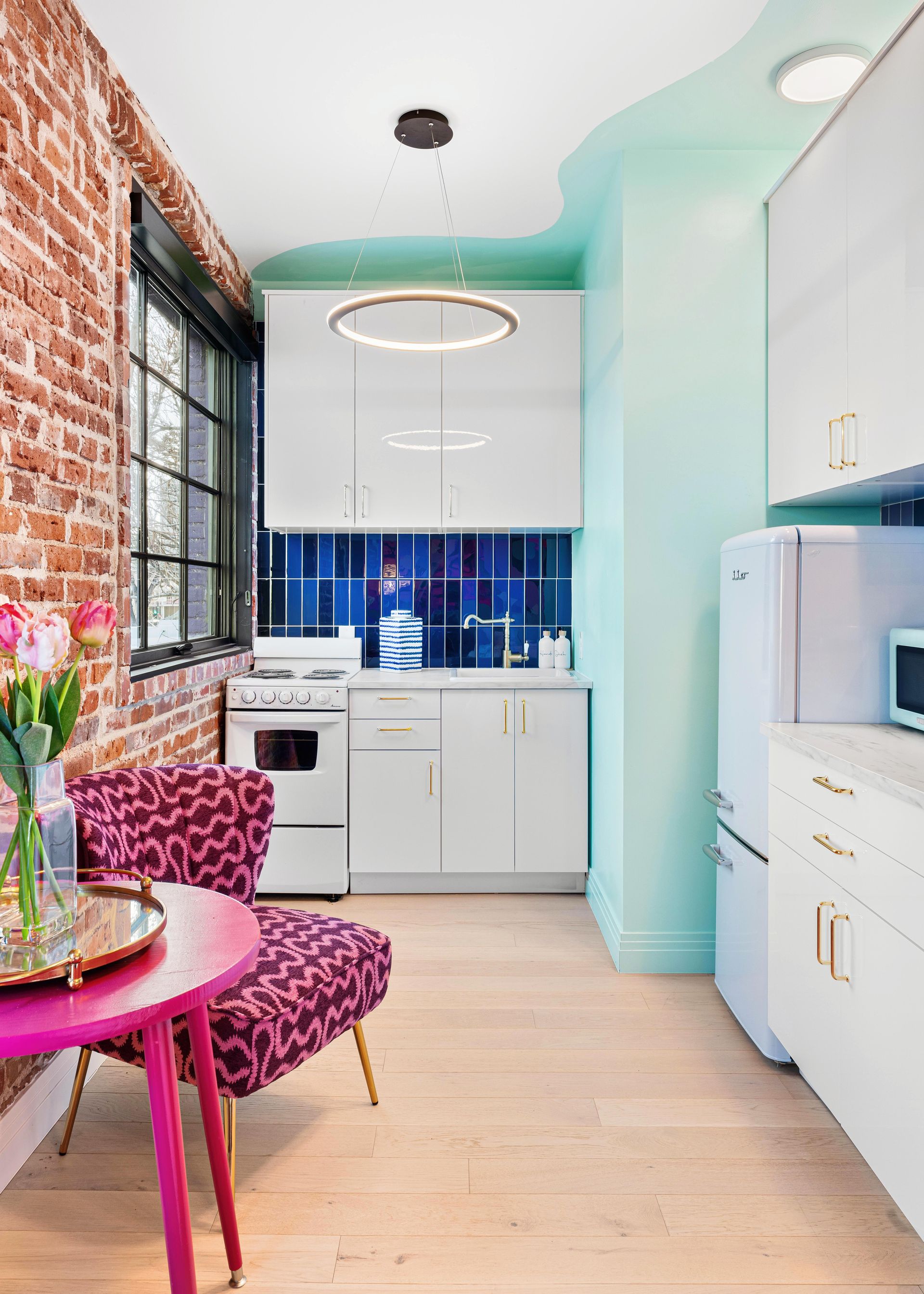 Narrow kitchen with mint walls, exposed brick, blue tile backsplash, white cabinets, and pink chairs