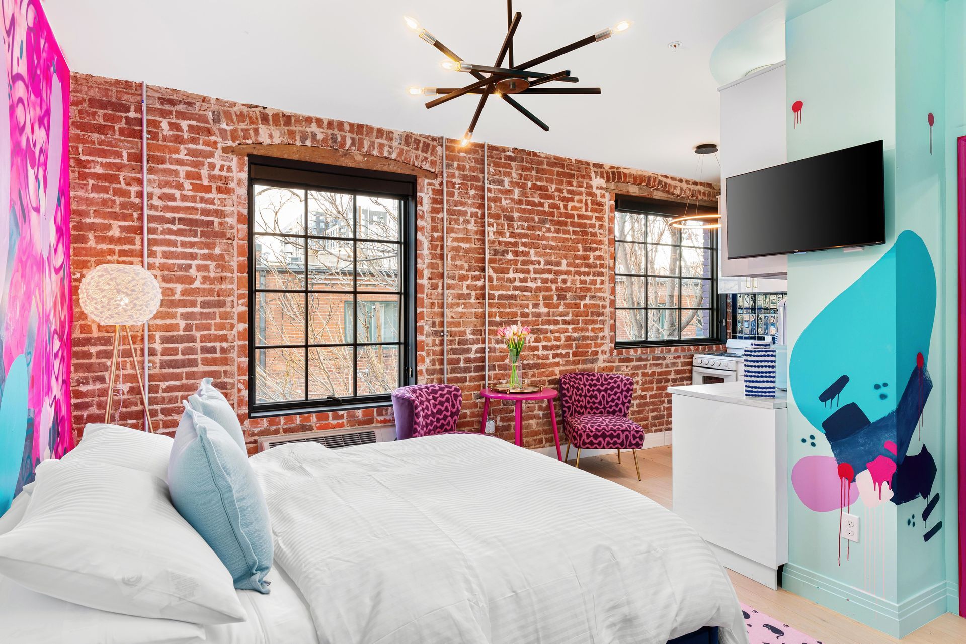 Bright bedroom with exposed brick walls, white bed, colorful chairs, and large windows overlooking a city skyline