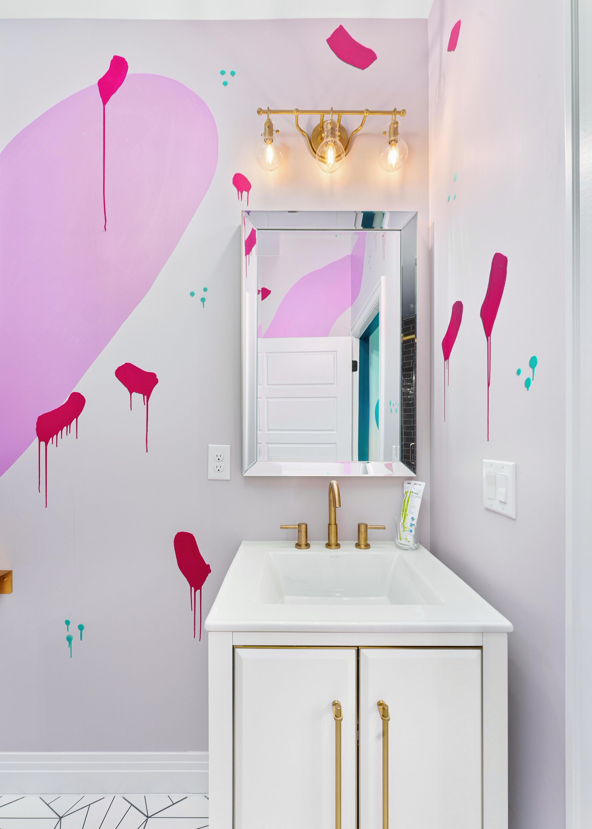 Small bathroom vanity with gold fixtures, mirror, and white walls accented by pink paint splatters.