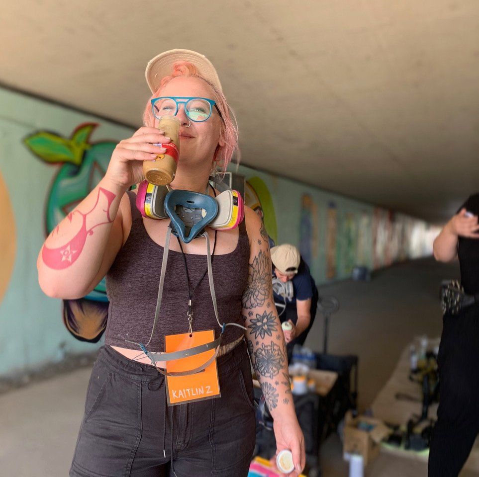 Person with pink hair and blue glasses drinks from a bottle in a graffiti-covered tunnel, wearing headphones.