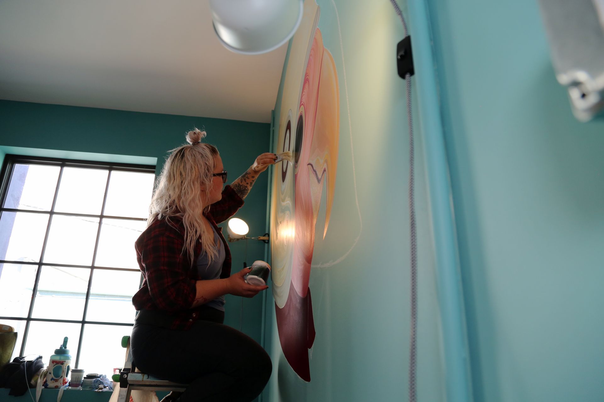 Person painting a large mural on a teal wall in a bright room with a window and ceiling light