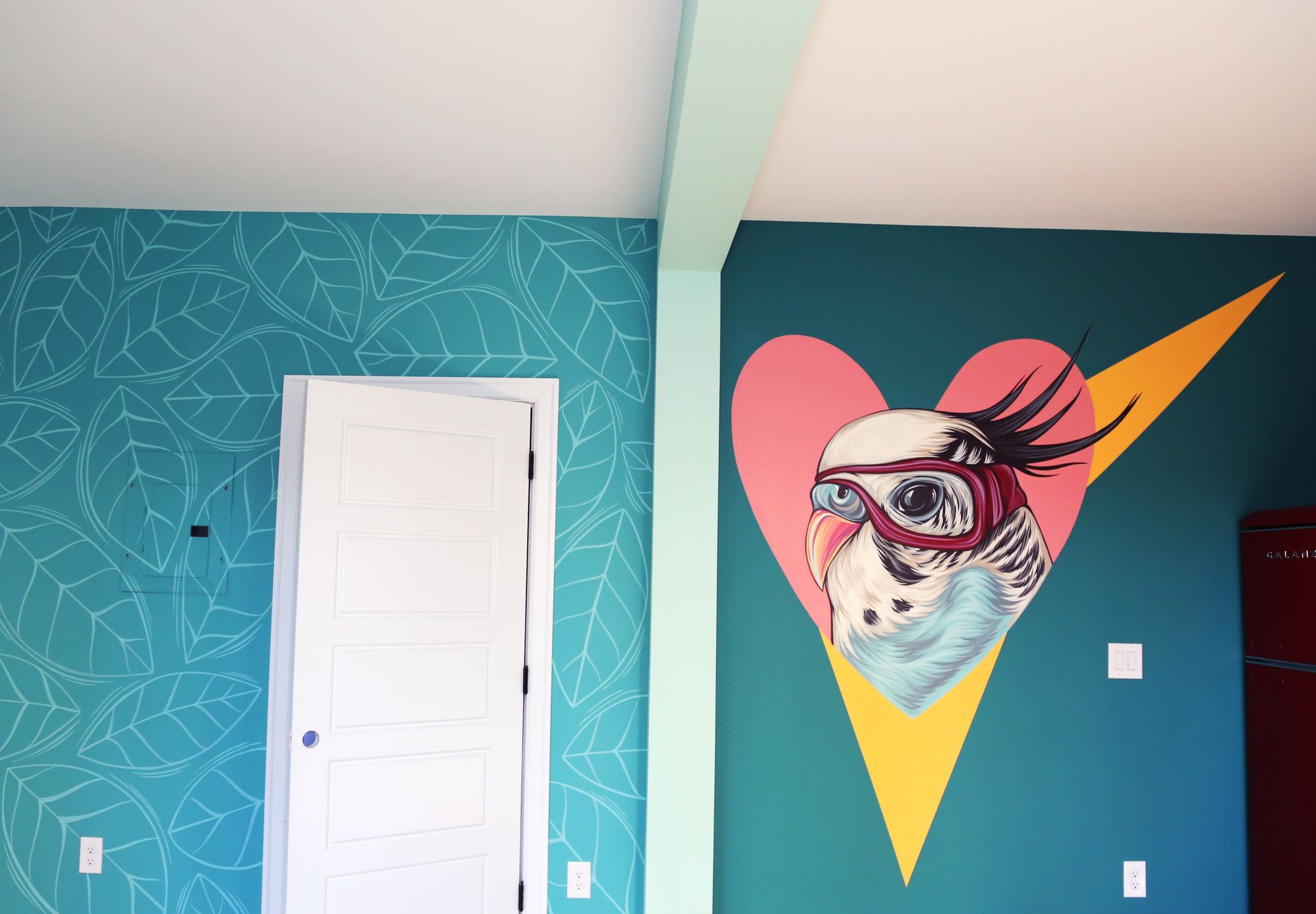 Teal room with a white door and a colorful wall mural of a heart, bird, and sunglasses