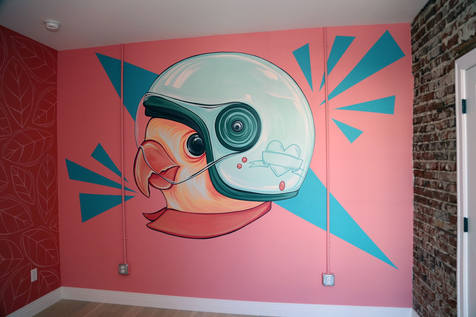 Pink wall mural of a bird head in a helmet with teal geometric accents in a room