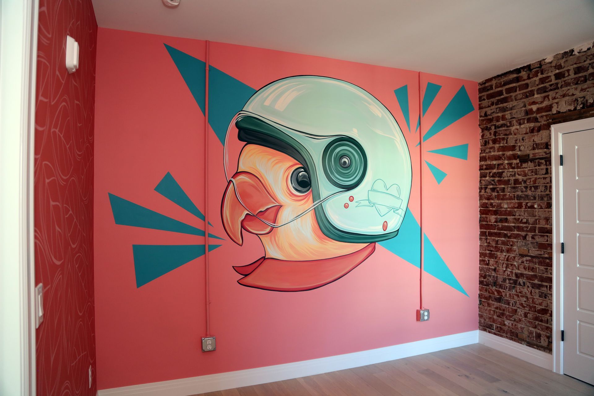 Mural of a helmeted bird on a pink wall with teal geometric shapes in a room with brick accents