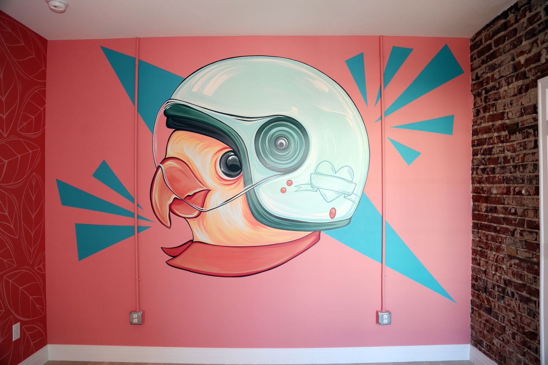 Mural of a bird in a helmet on a pink wall with teal geometric accents and a brick edge