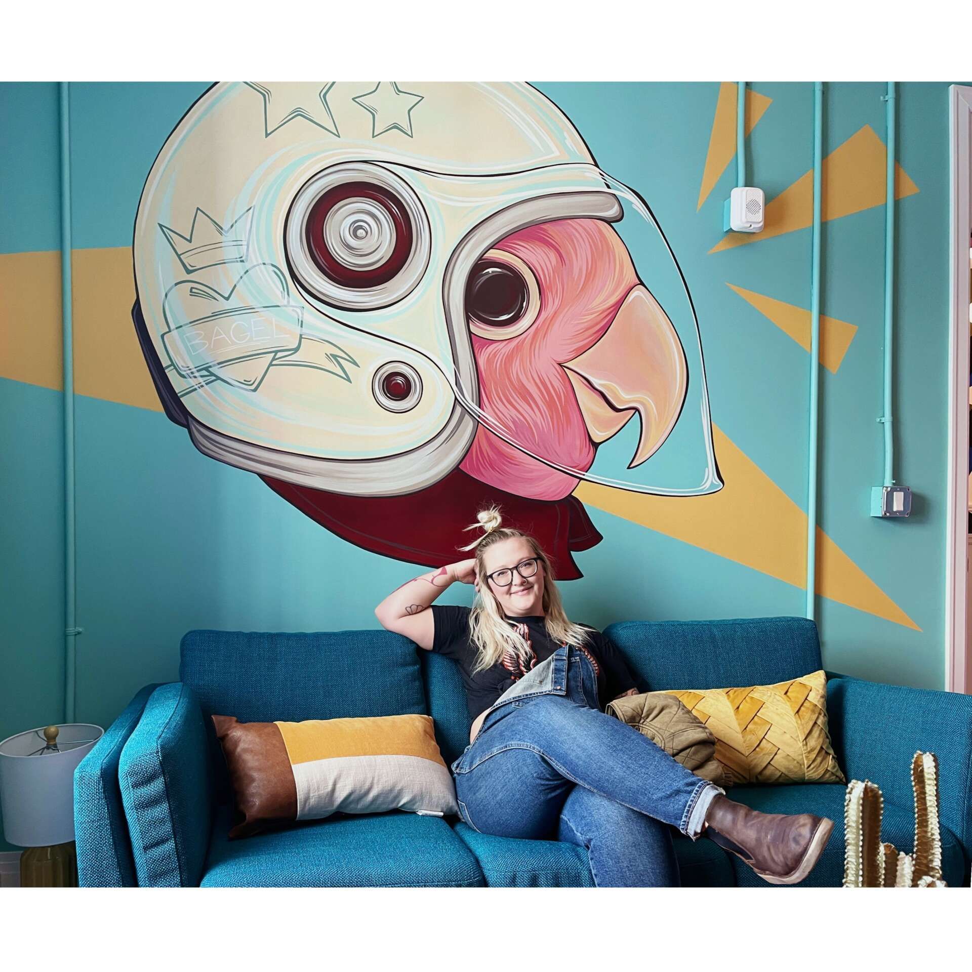 Person lounging on a blue couch beneath a colorful bird mural in a bright room